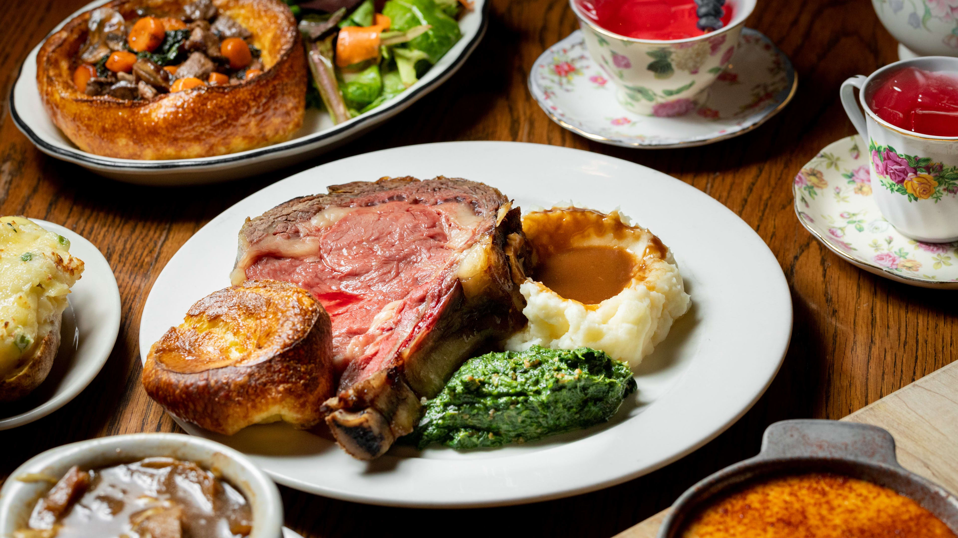 A spread of dishes including a steak with mashed potatoes, meat pie, cocktails in teacups and soup.