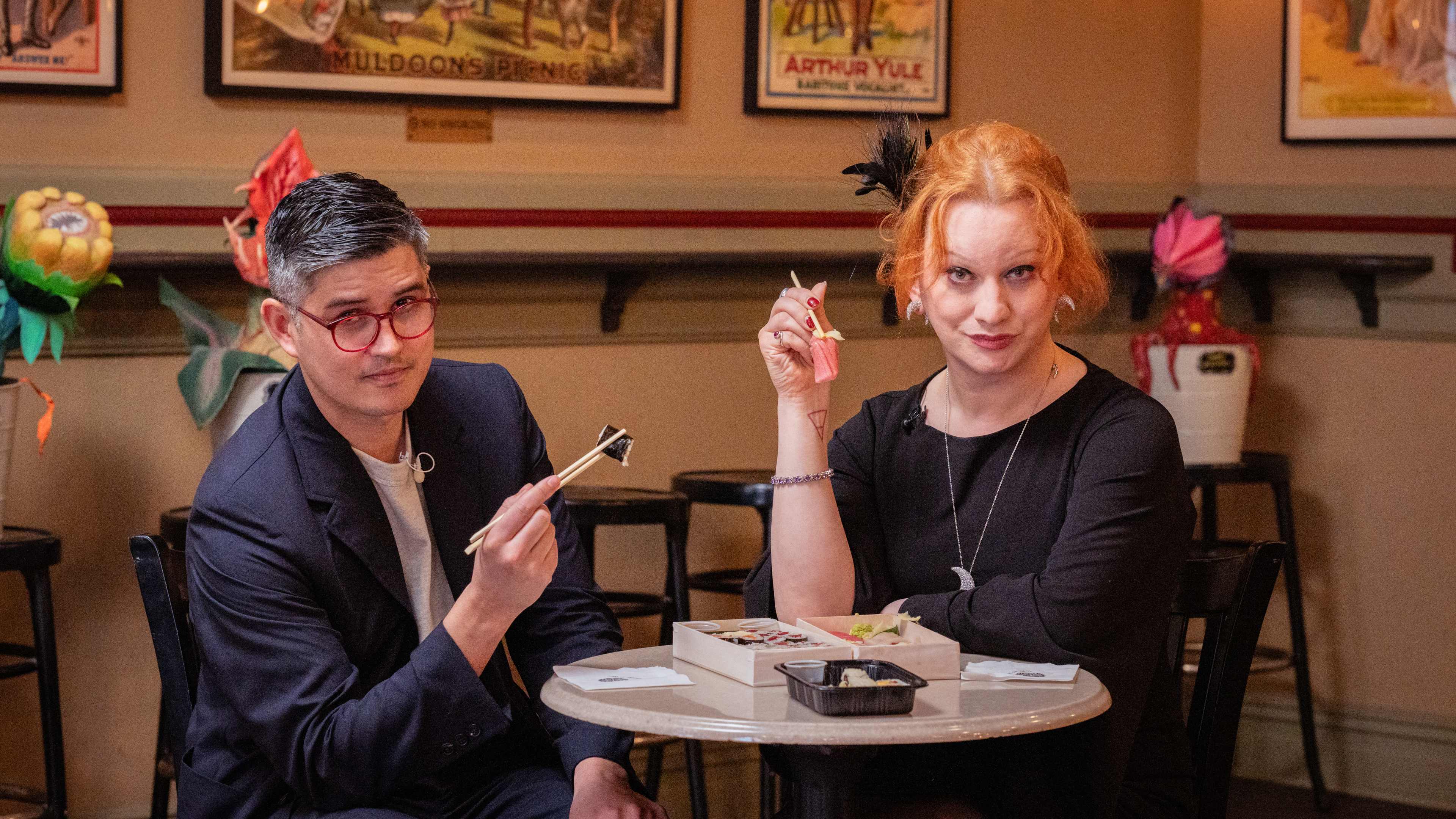 two people sit at a table. they are both holding sushi in chopsticks held out in front of them