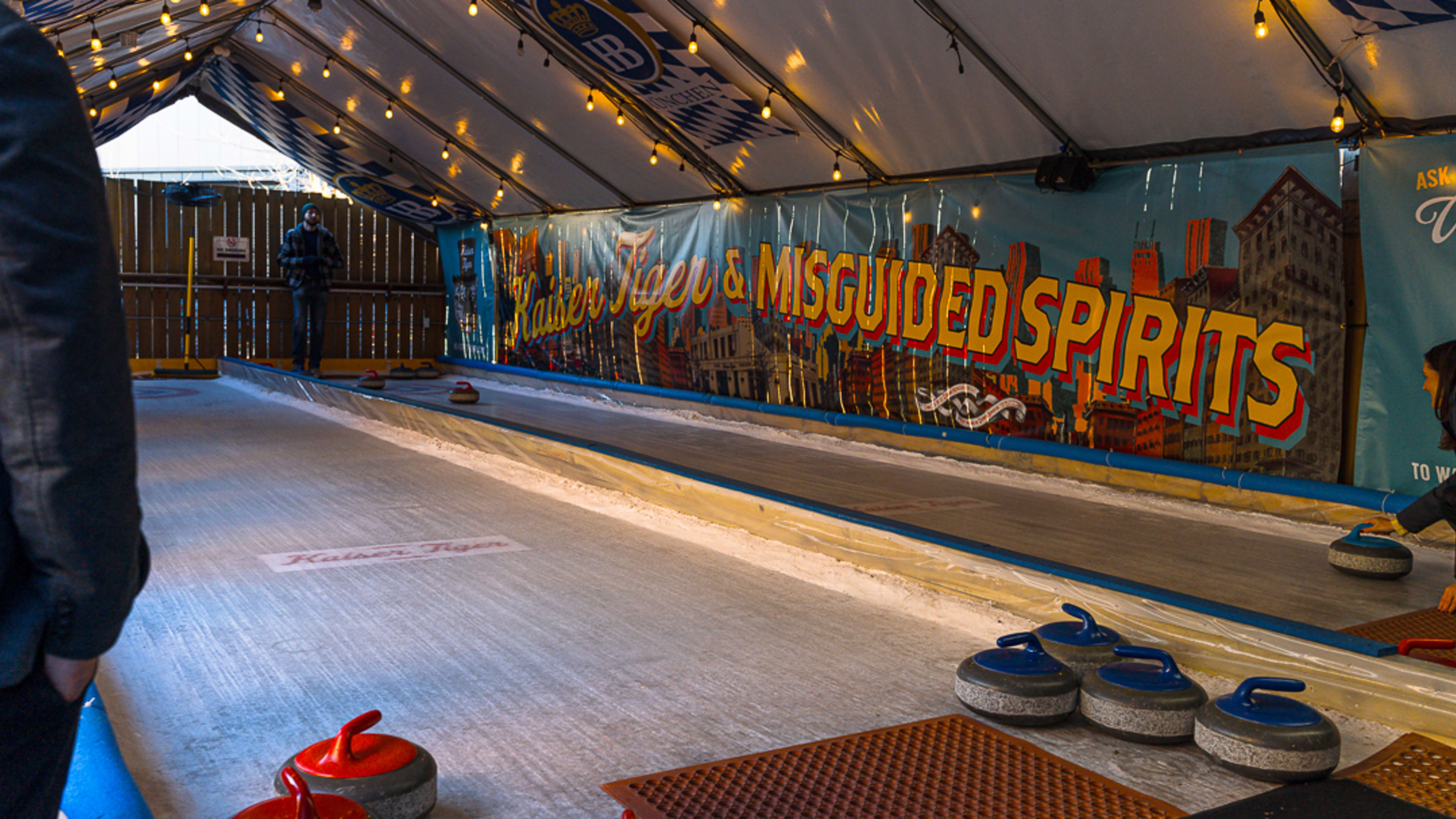 A curling rink in an enclosed tent with hanging twinkle lights and a banner that reads, "Kaiser Tiger & Misguided Spirits."