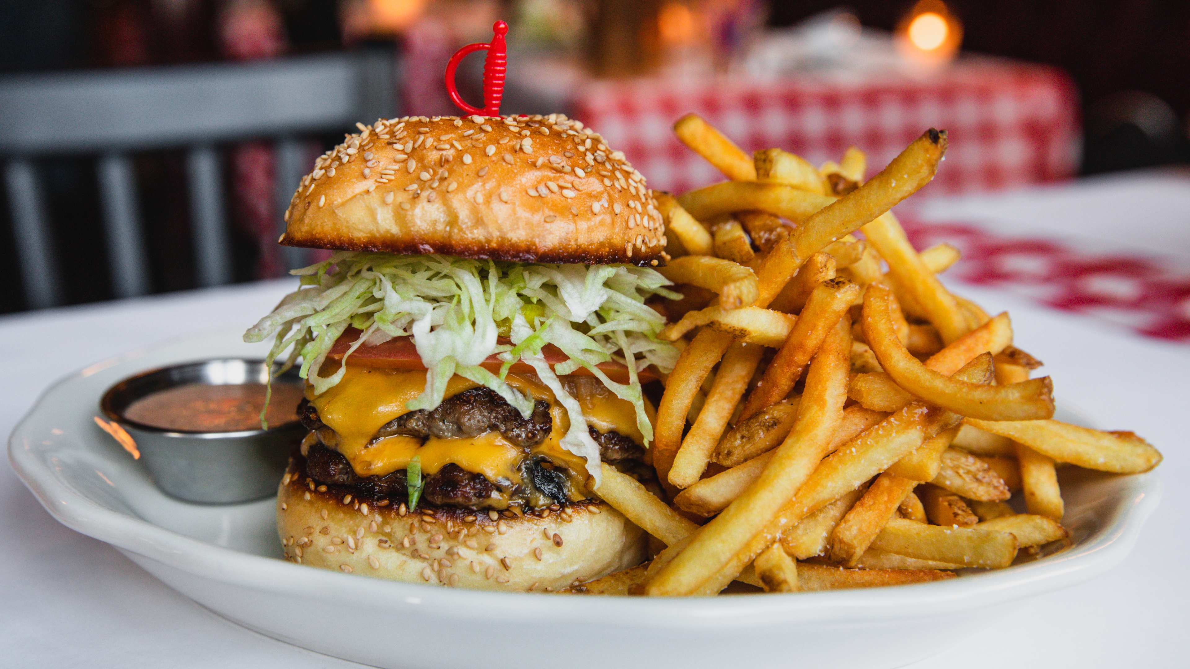 A double cheeseburger on a white plate with a side of fries and sauce. There is a cocktail sword stuck through the center of the burger.