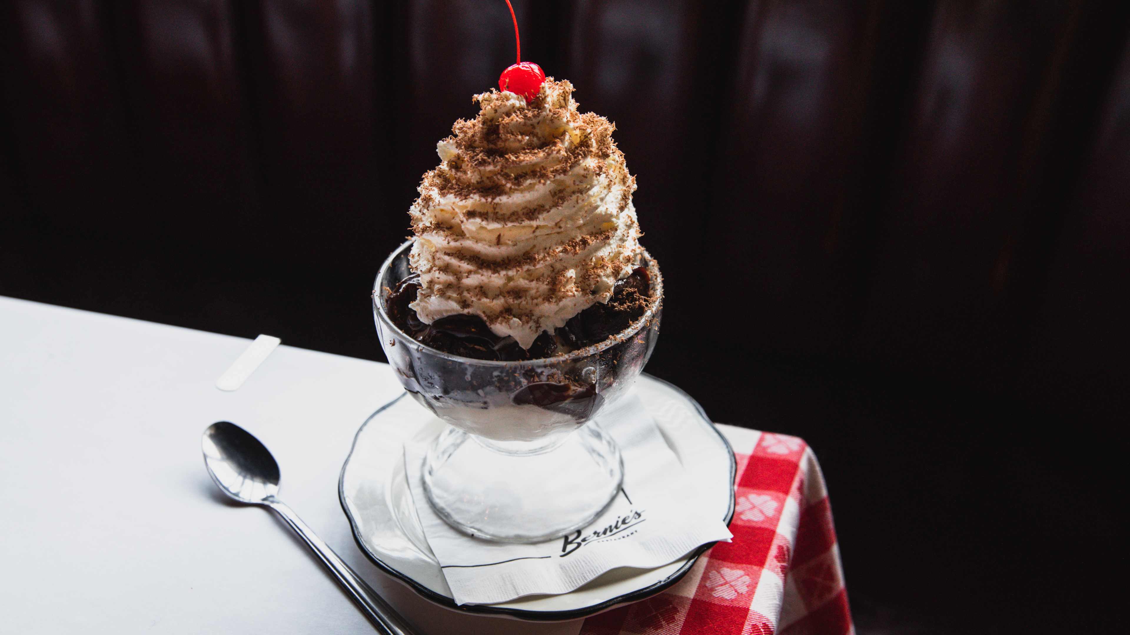 The Brownie Sundae from Bernie's in a glass sundae cup, on a white plate, with an ice cream spoon on the site.