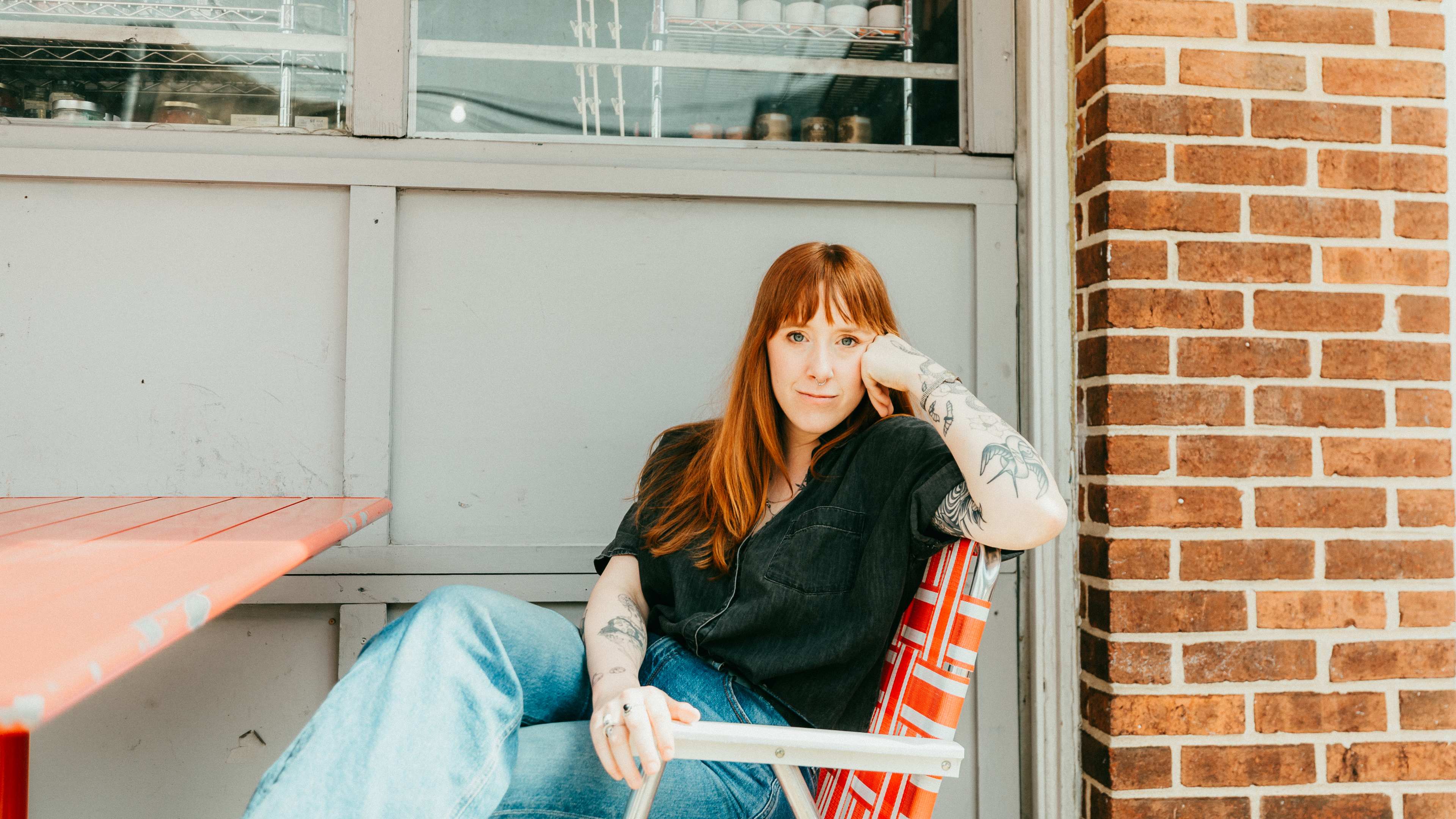 a woman with auburn hair wears a black top and blue jeans while sitting in a red chair