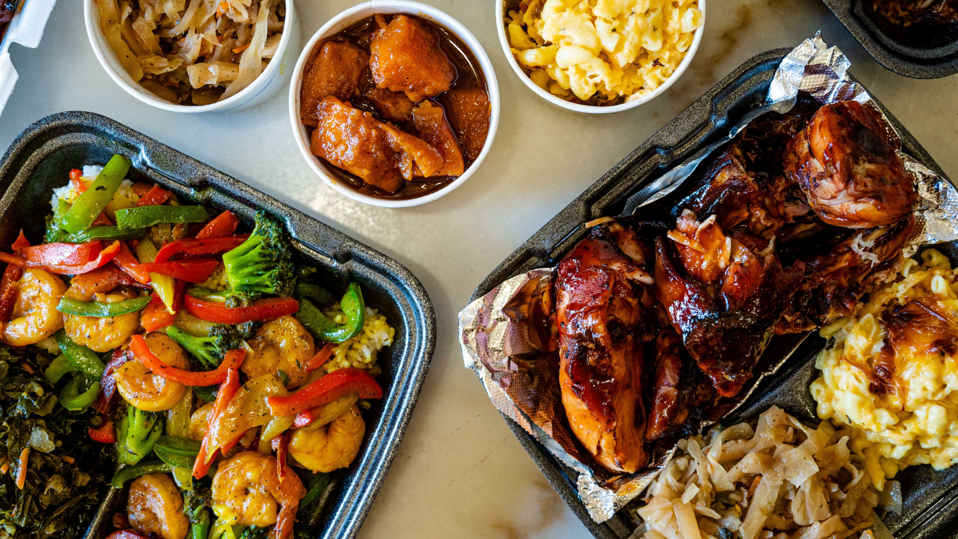 spread of takeout plates of wings, shrimp, veggies, mac and cheese, and yams