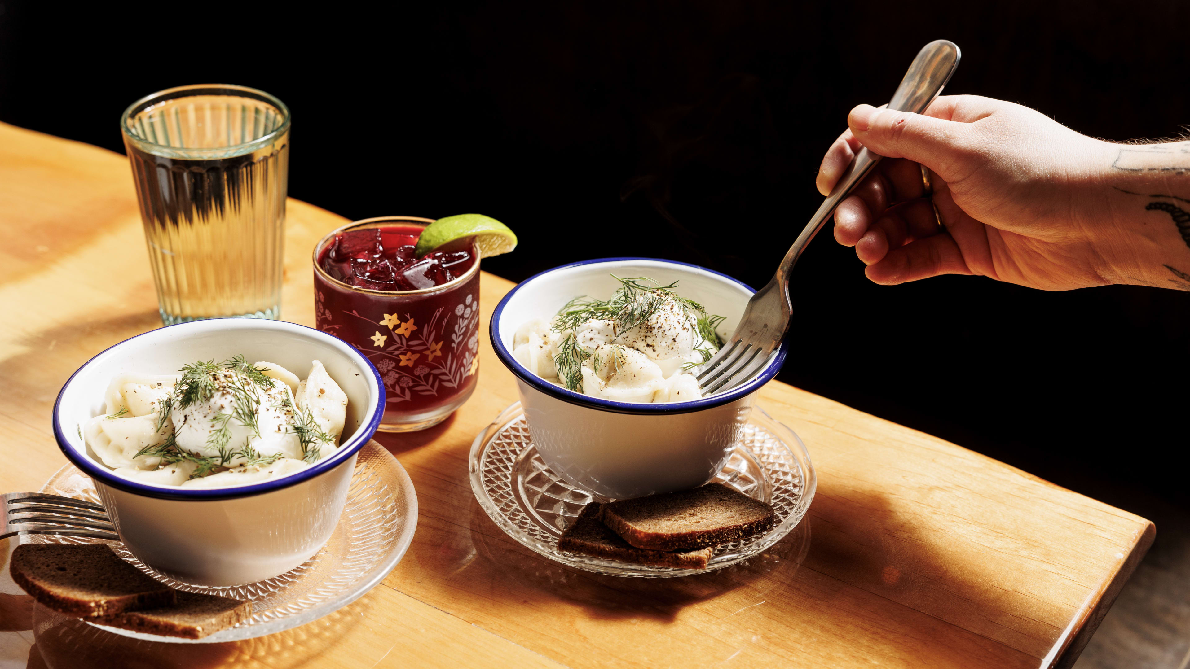 Bowls of pelmeni with a cocktail in a short glass and a fork reaching into the bowl.