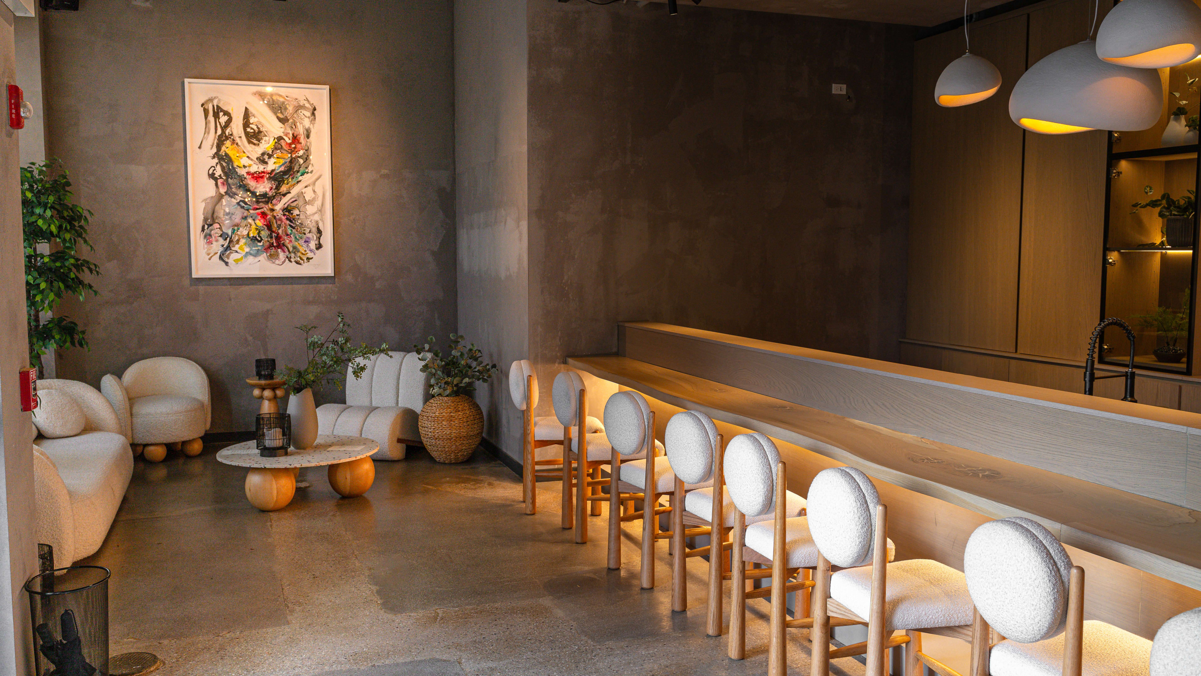 The grey slate interior of Kyoten Next Door. The sushi bar is to the right, lined with white chairs.  There are white ovular hanging pendants. There is a seating area in the back corner with plants, white mod furniture and a low white coffee table.