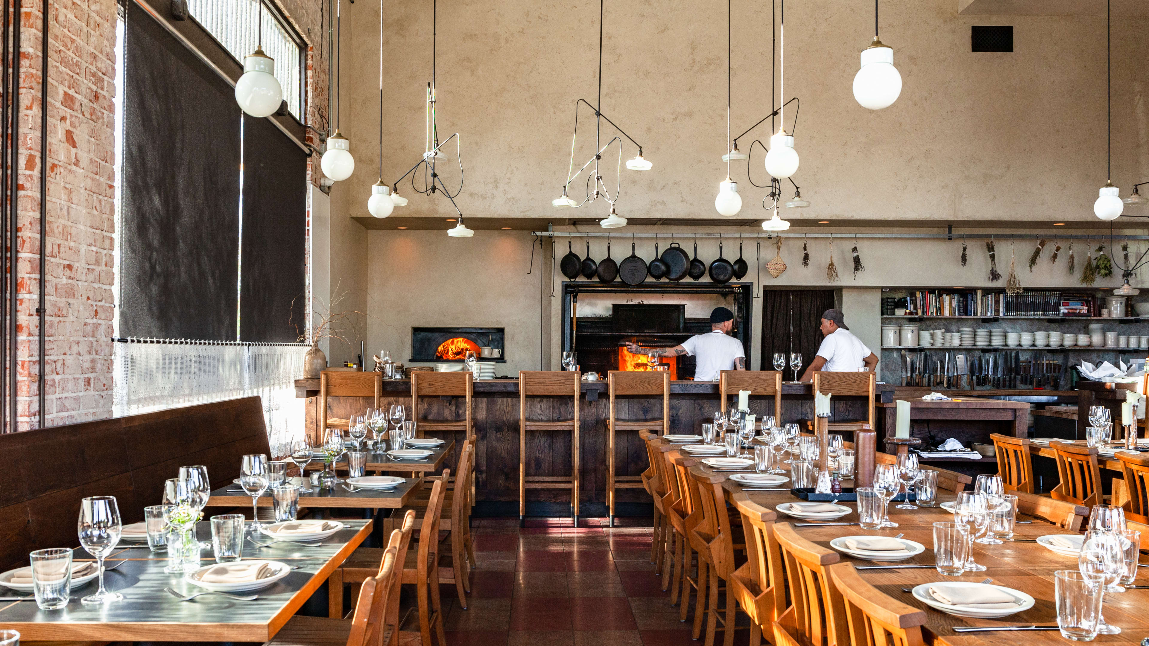 The interior of the dining space at Dunsmoor. There are wooden tables, high ceilings, hanging bulb pendants, and cast iron pans hanging above a live fire hearth.