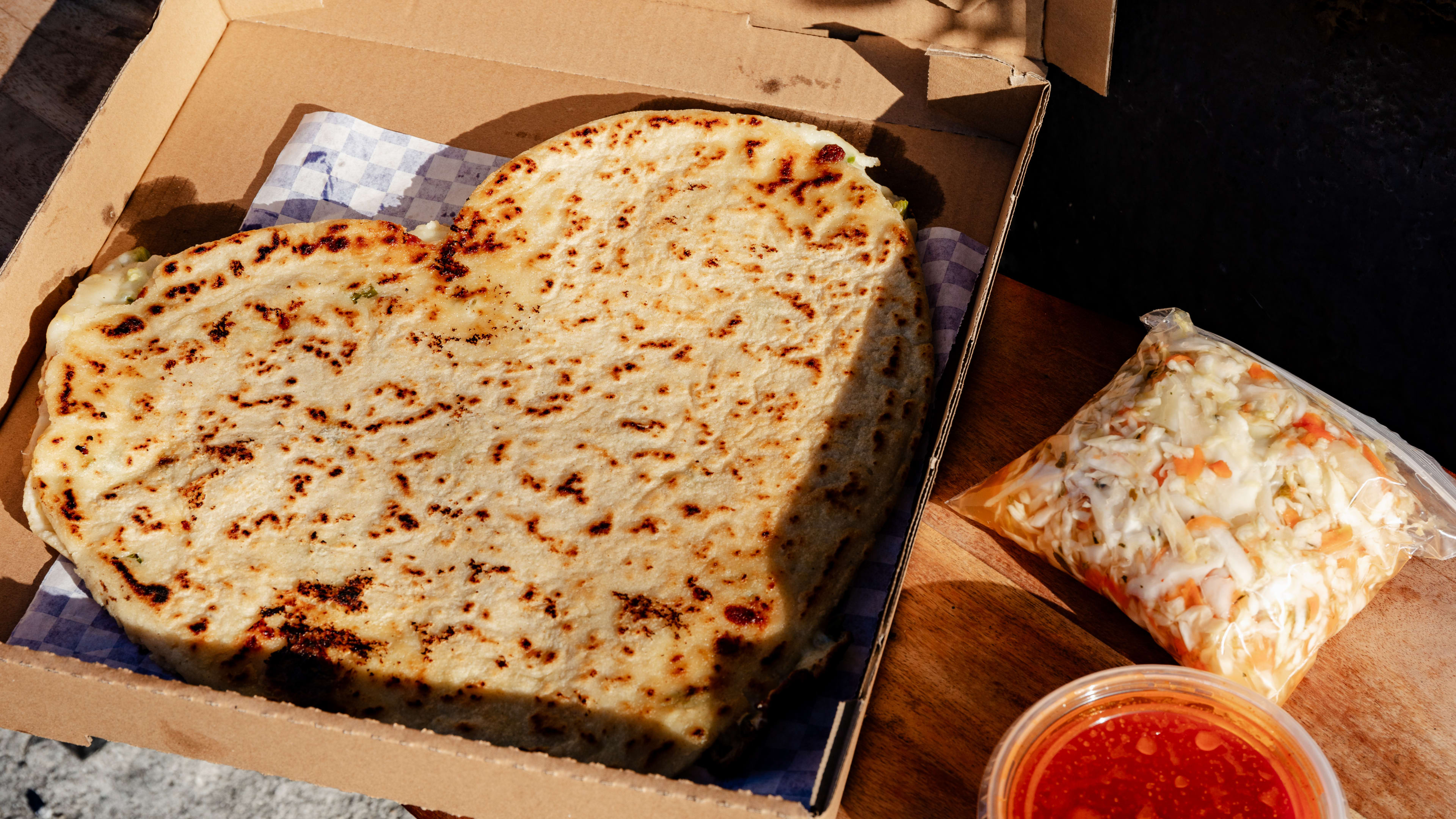 A heart shaped pupusa in a brown pizza box with toppings on the side.