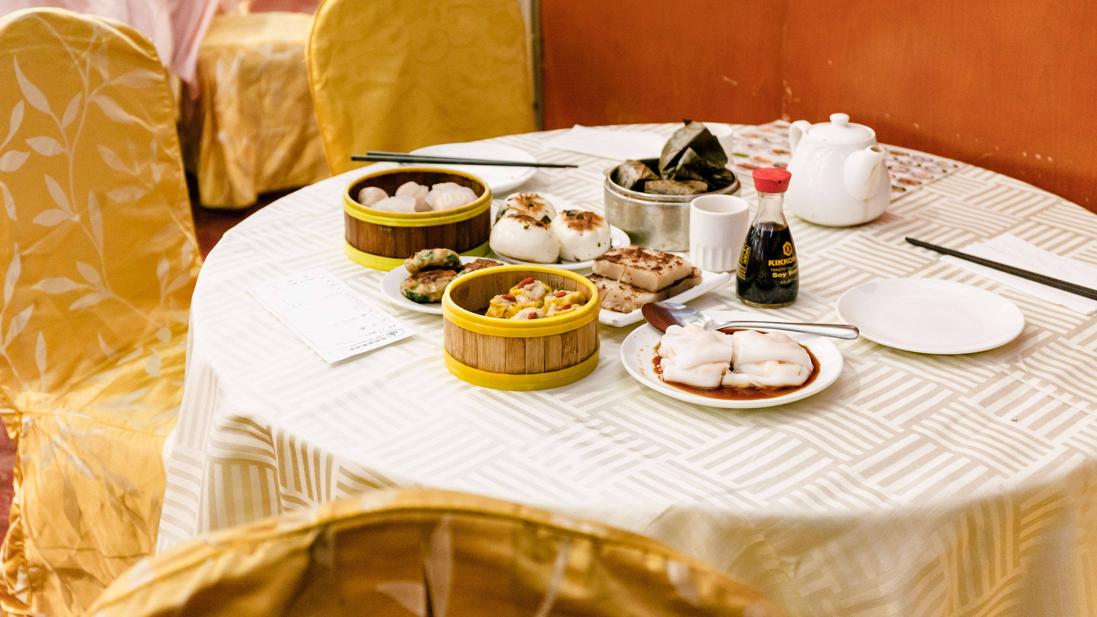 A spread of dishes on plates and in steamer baskets on a white clothed round table with gold fabric covered chairs.