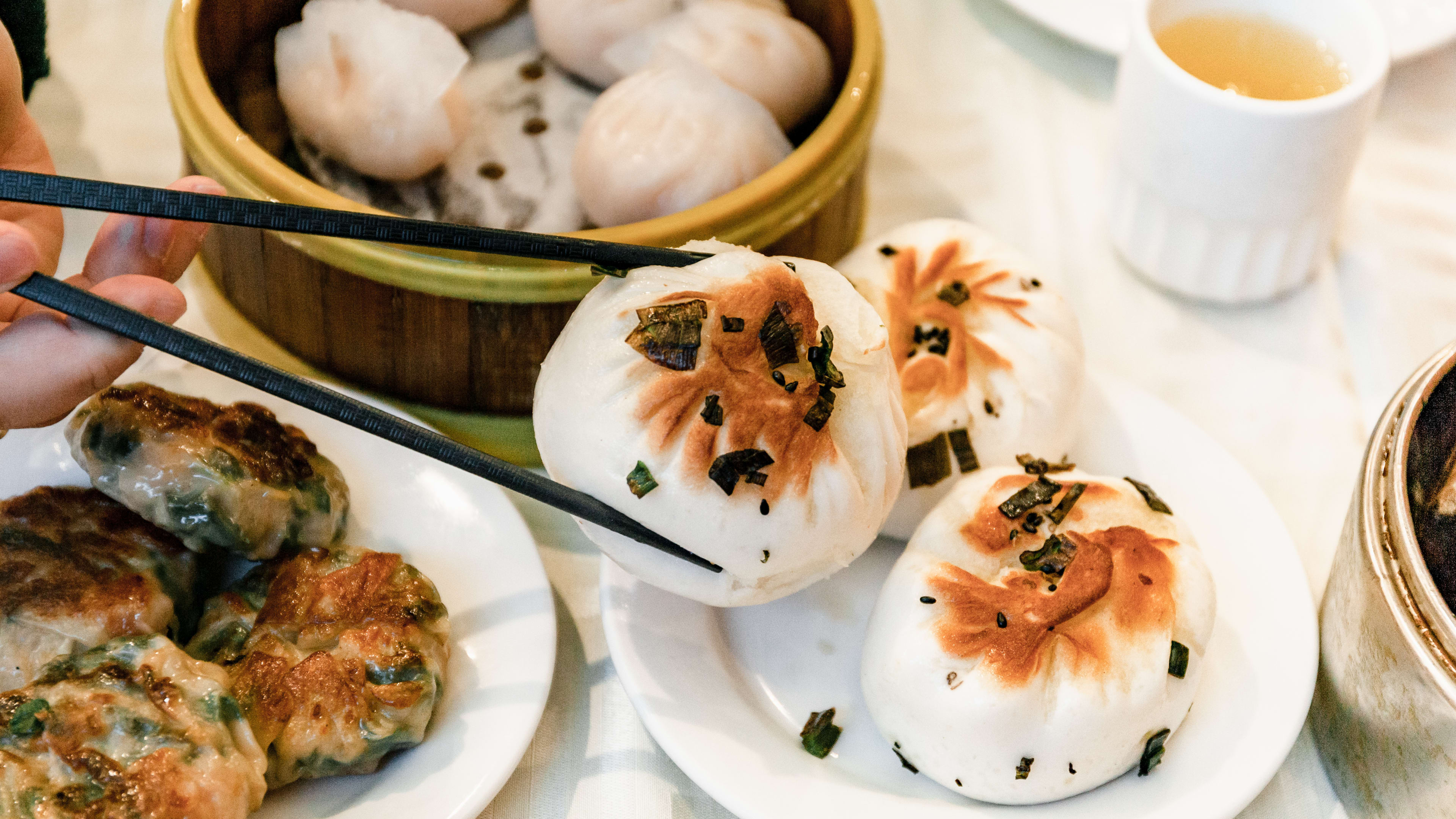 Dumplings in a steamer basket, meatballs, and a person holding up a bao with chopsticks from a plate.