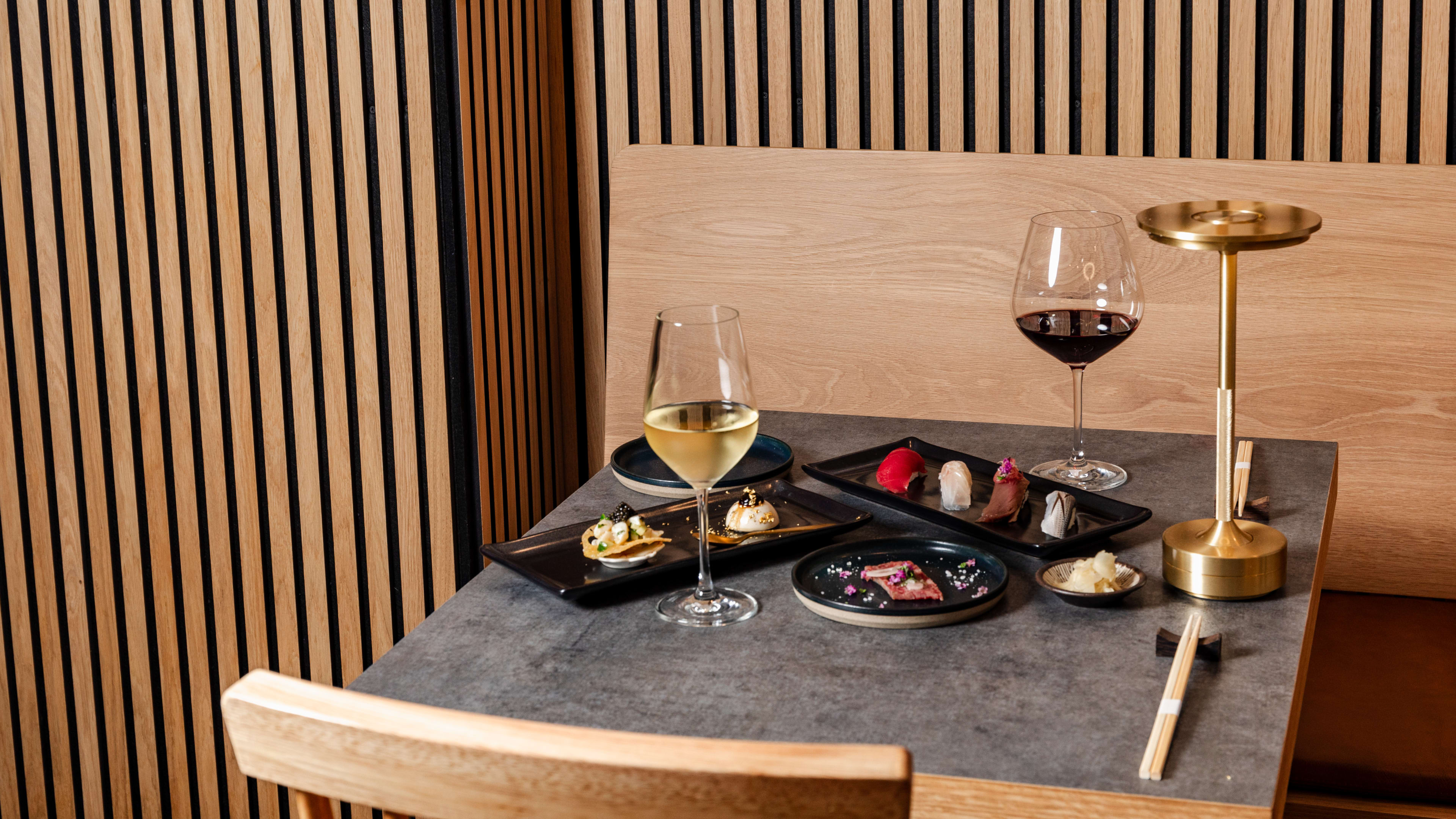 A spread of dishes on slate dish ware, glasses of wine, and a gold lamp on a grey wooden table, with wooden seating and slated wood walls.