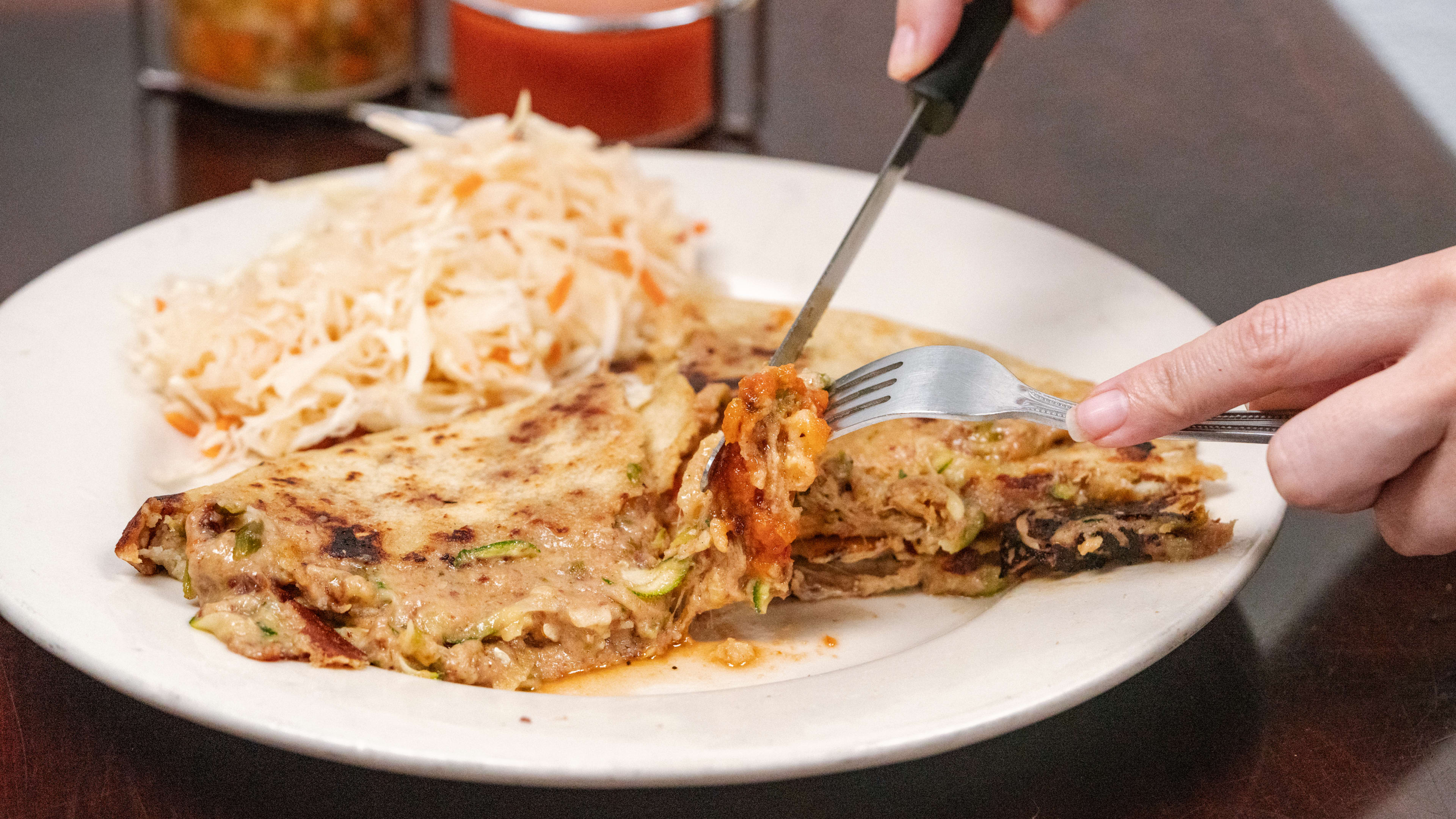 Large pupusa with hefty serving of cabbage on the side and a knife and fork cutting through it.