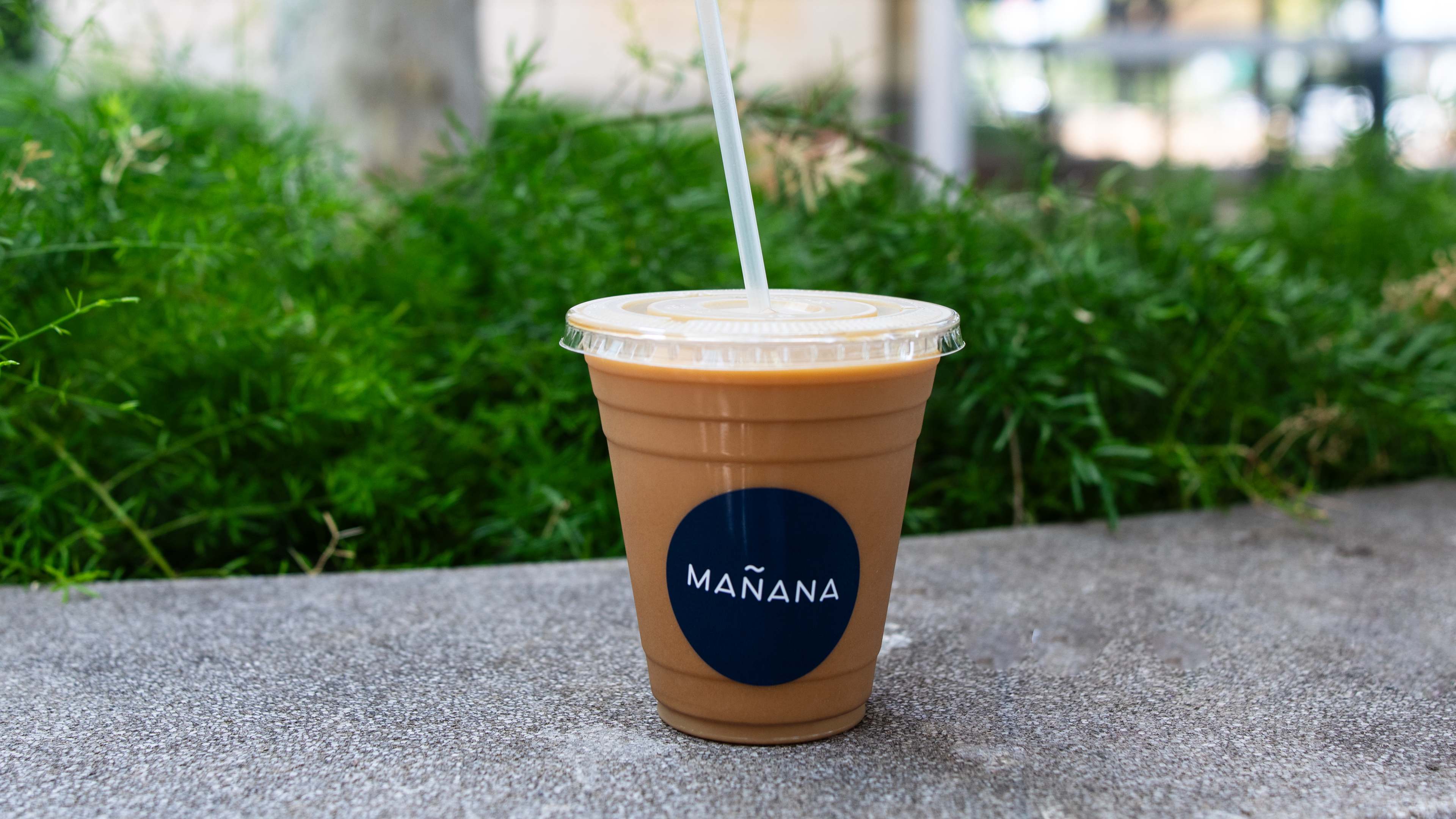An iced coffee drink in a plastic cup with a Mañana branded sticker, sitting on a concrete ledge outside.