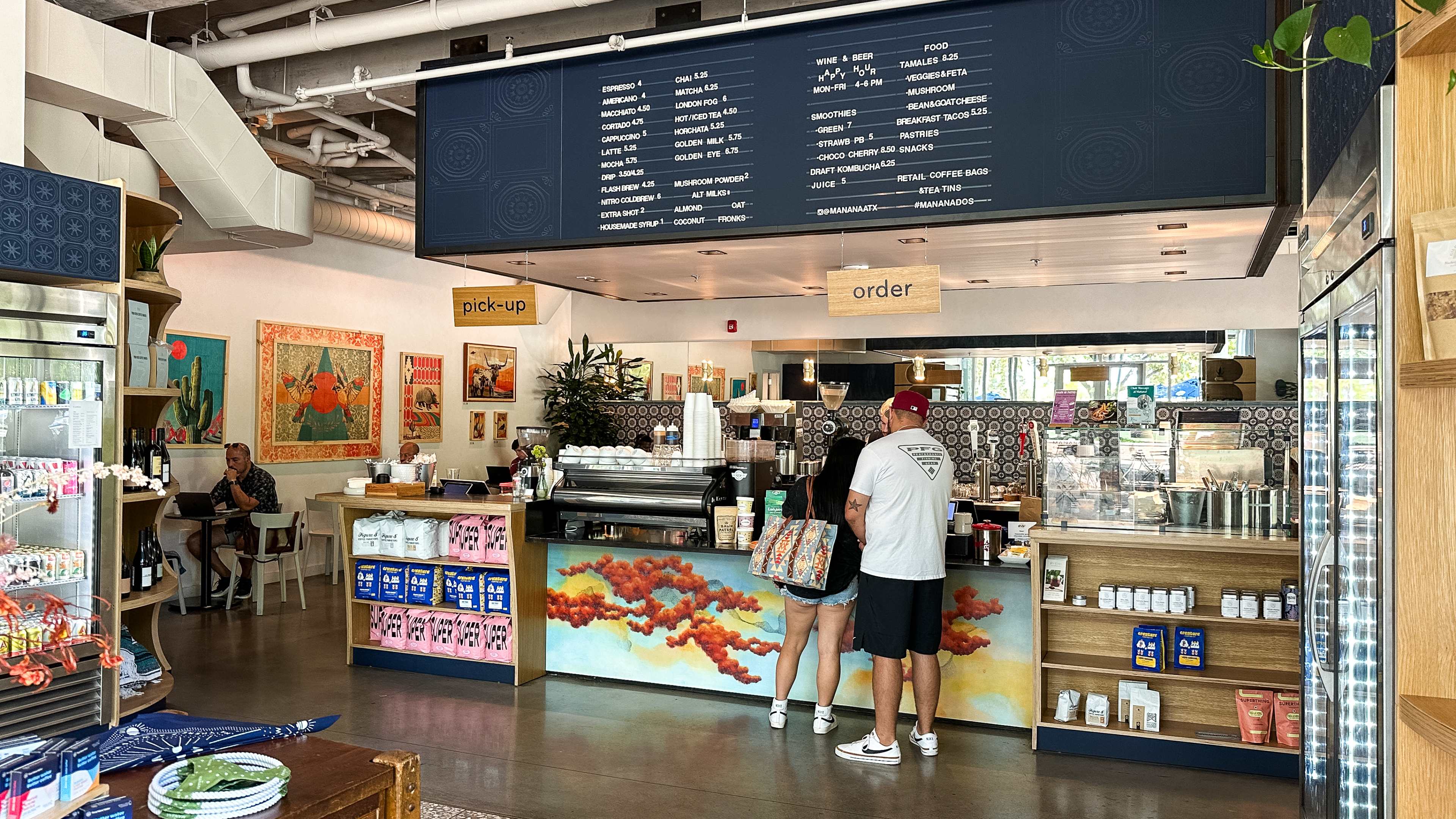 The interior of Mañana with grocery items on a table, shelves and in a refrigerator. There are people at a counter ordering.