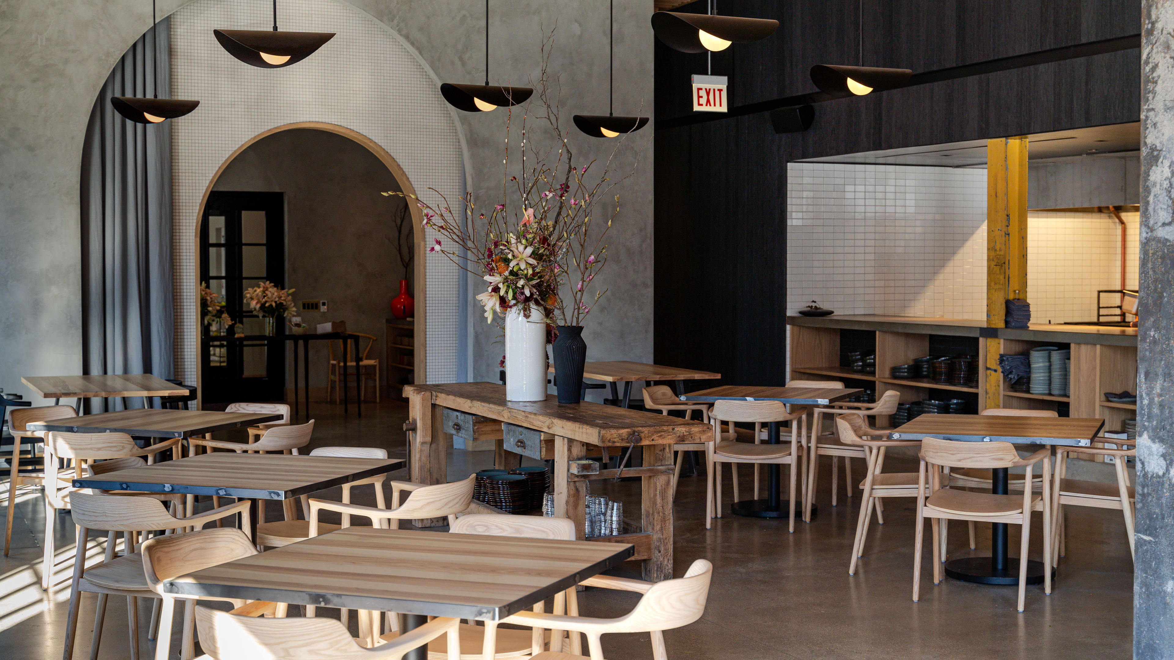 A neutral-toned dining room full of light-colored wooden tables and chairs, with modern light fixtures hanging from the ceiling.