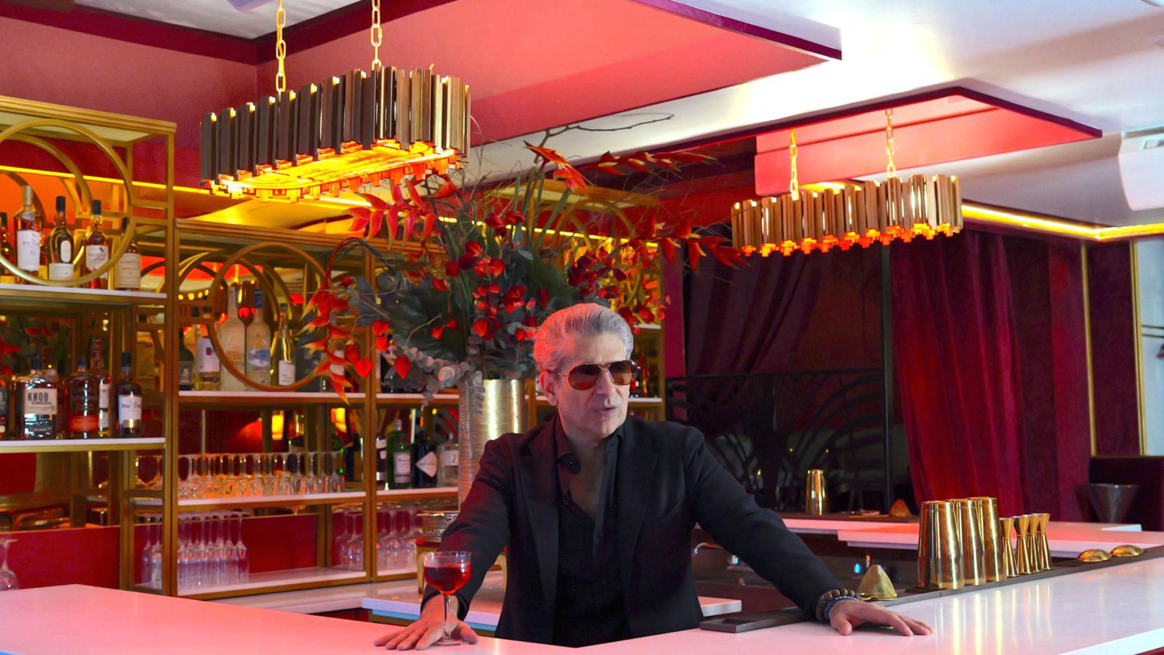 the actor michael imperioli wears sunglasses and a black jacket and stands behind a bar decked out with gold tumblers and accents