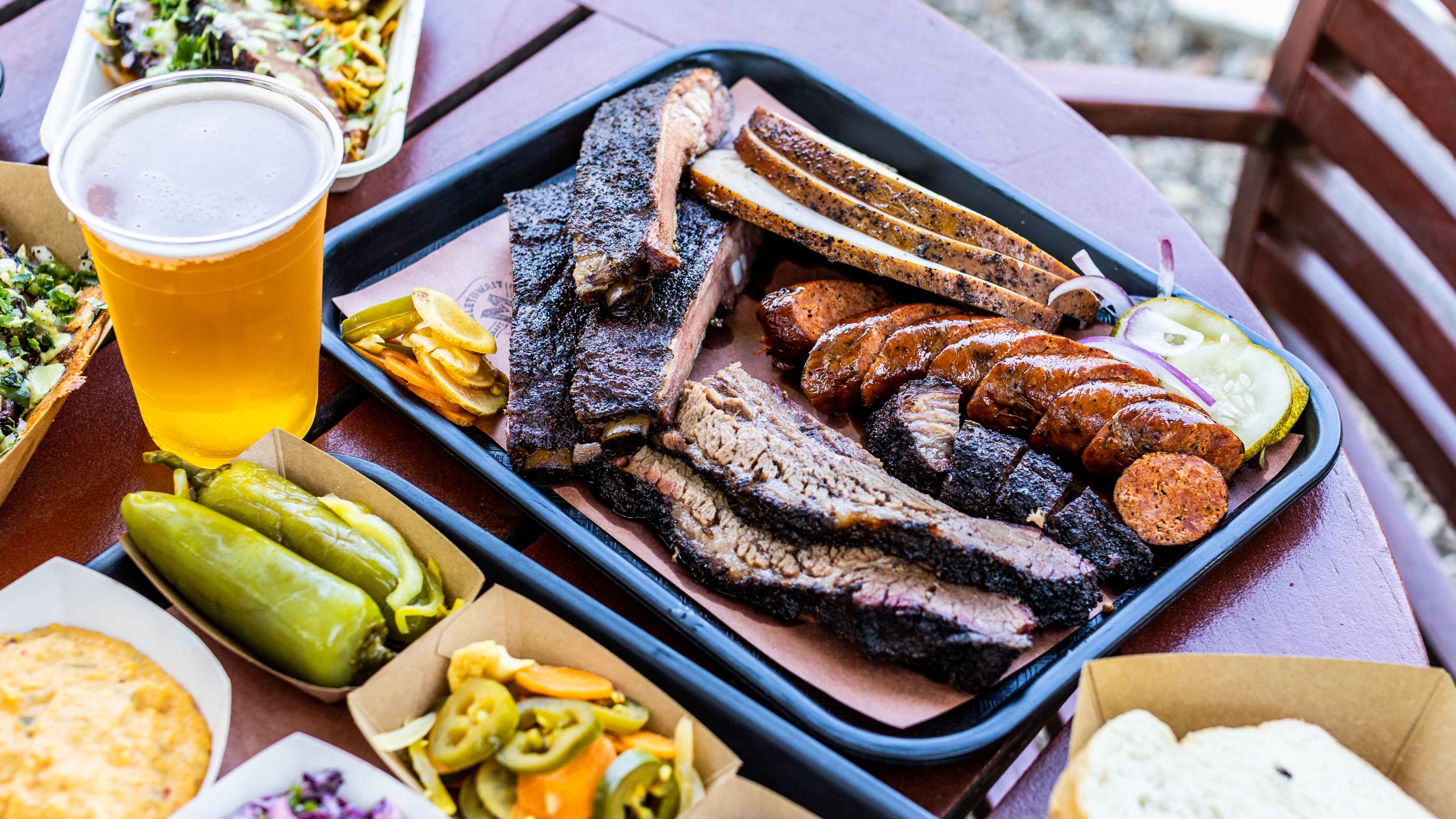 The BBQ Platter at Micklethwait, served on a black lunch tray. There is a tray of sides in brown takeway bowls, and a plastic cup of beer.