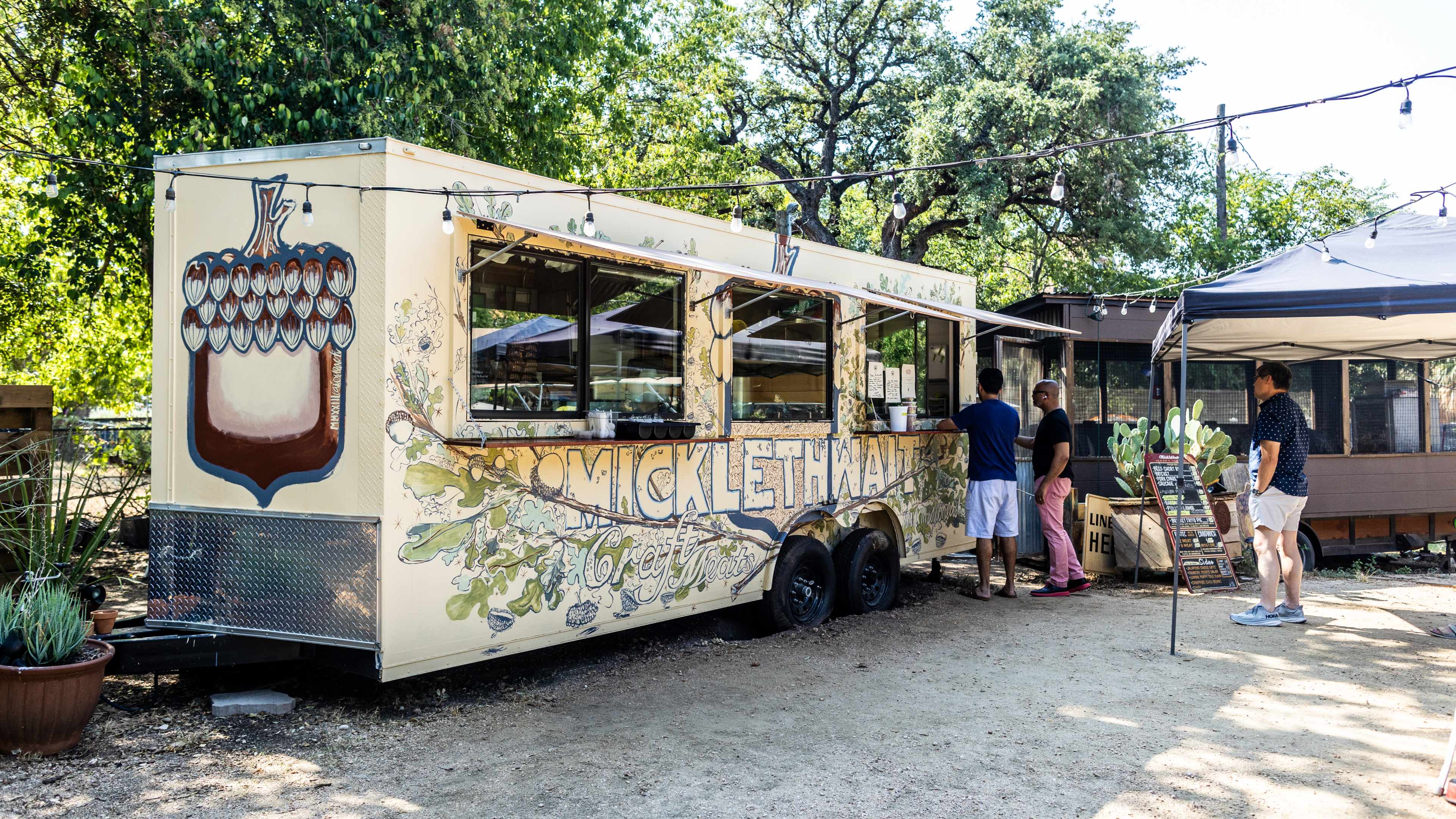 The food truck exterior of Micklethwait. There are people standing in line, hanging string lights, a tent and potted plants.