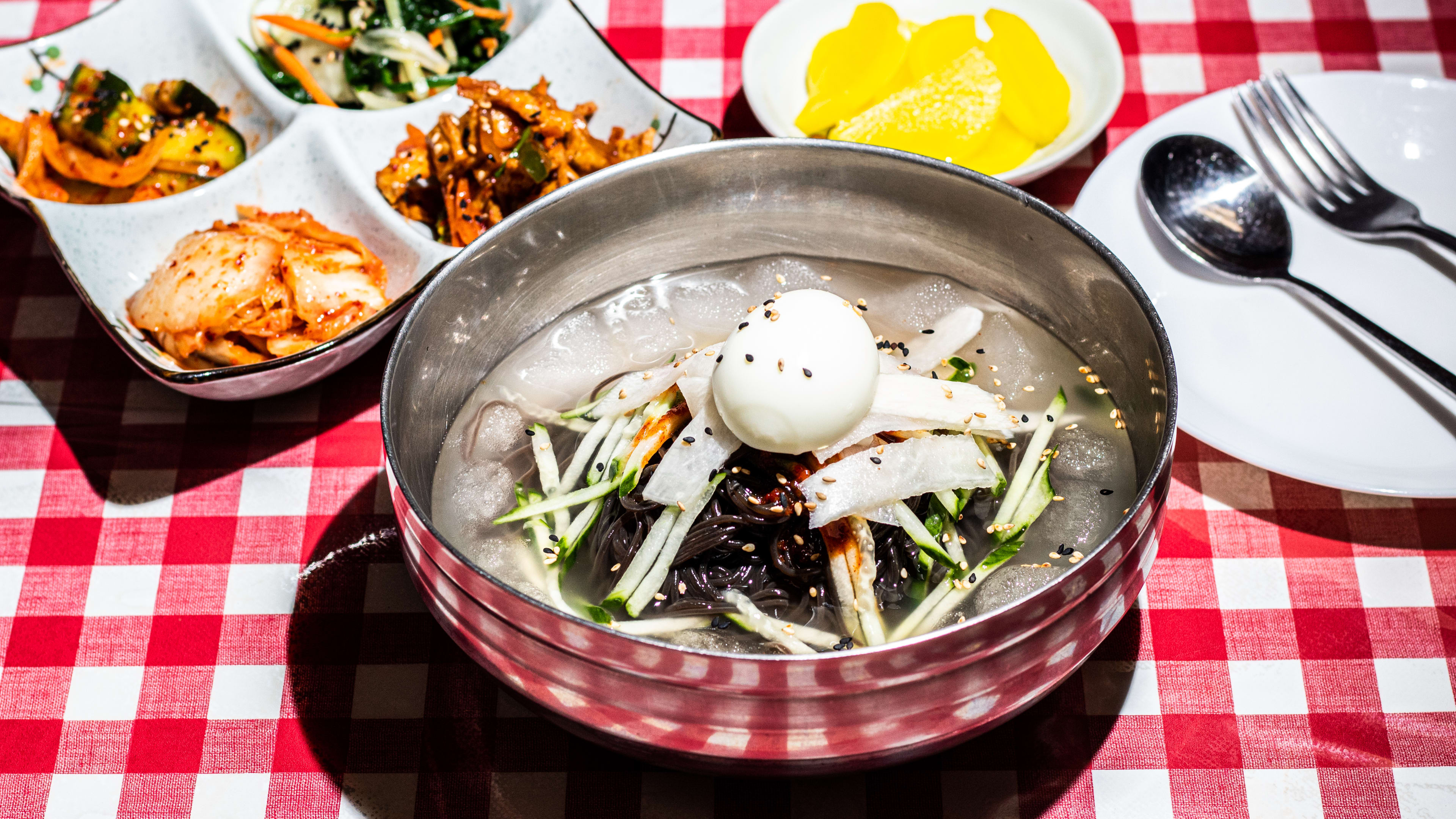 Spread of dishes including kimchi/banchan, cold noodle dish in tin bowl with whole egg, and cutlery.