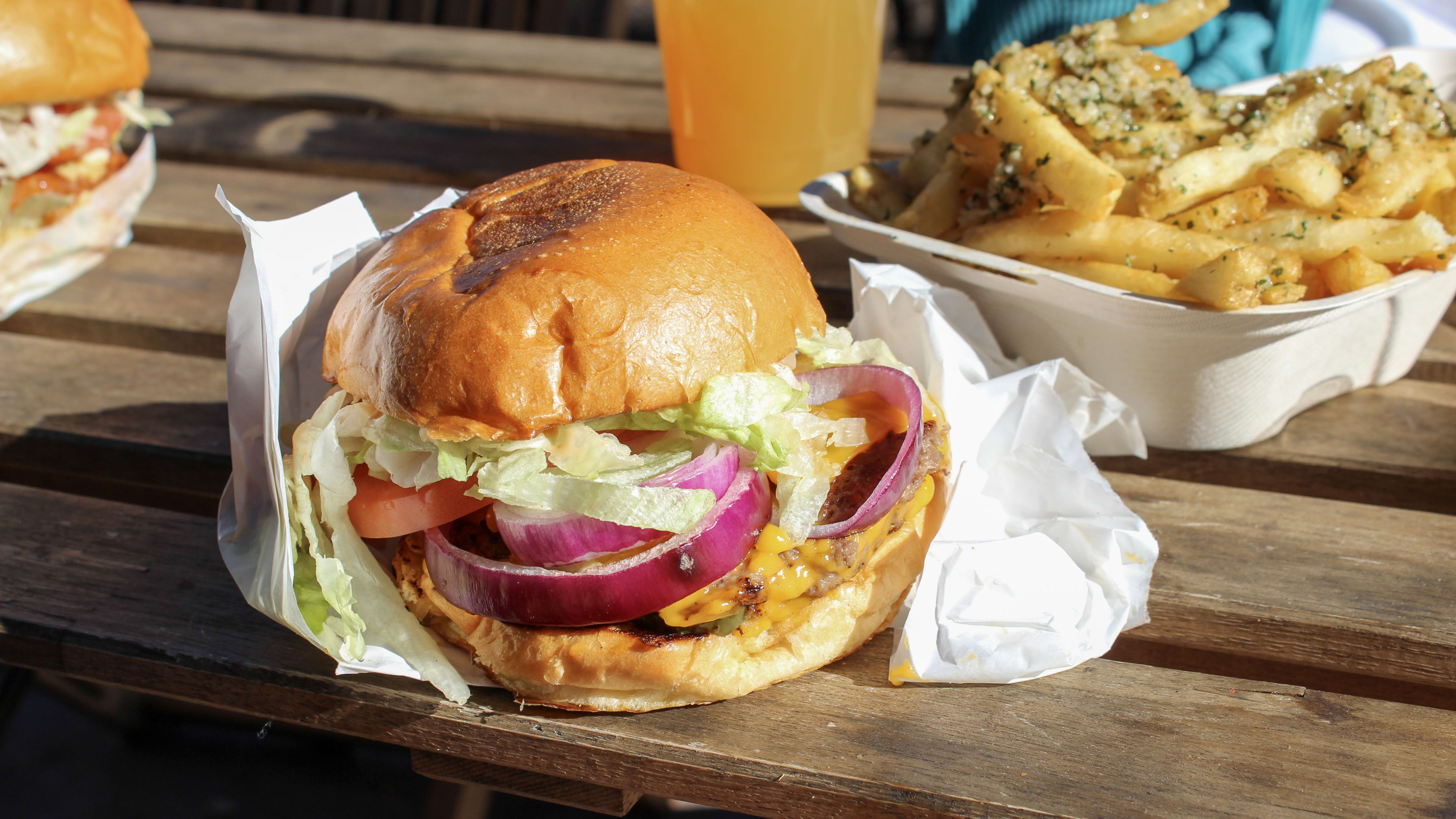 A burger wrapped in paper sits on a wooden table with fries and a beer in the background.