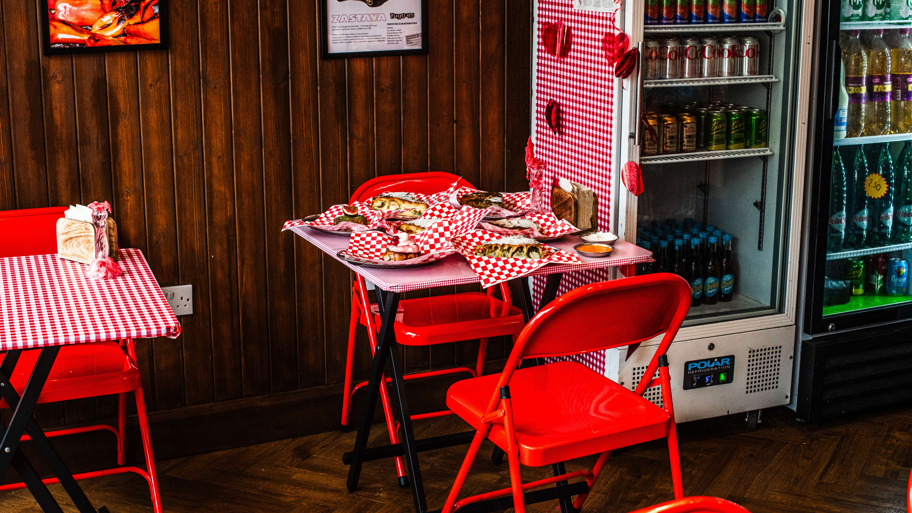 The interior of Mystic Burek with wood paneled walls, two refrigerators with drinks, and two tables with red and white checkered table cloths, red folding chairs, and various burek slices on one table on top of red and white paper.