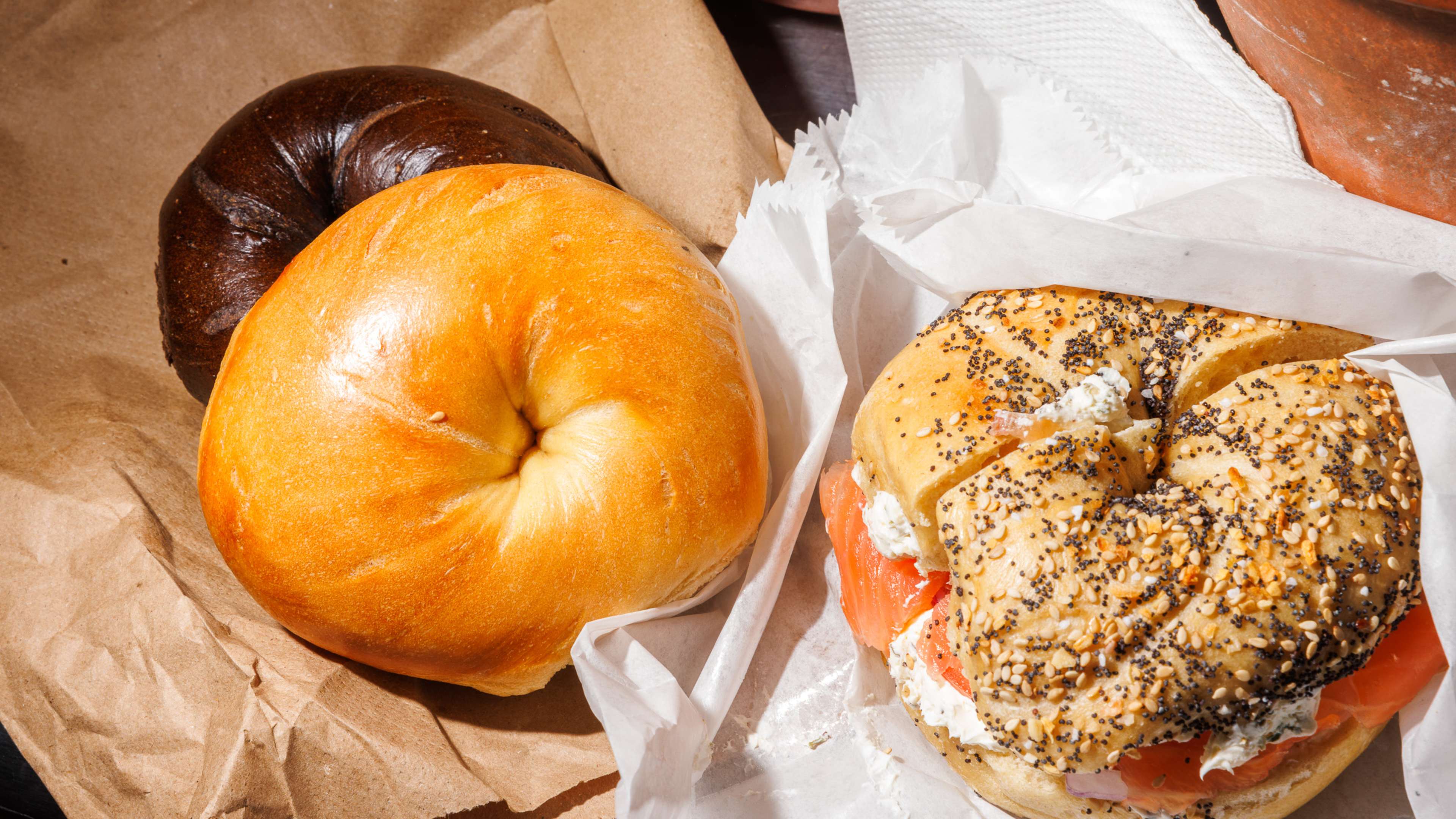 A pumpernickel and plain bagel on top of a brown bag next to an everything bagel with cream cheese and lox on white paper.