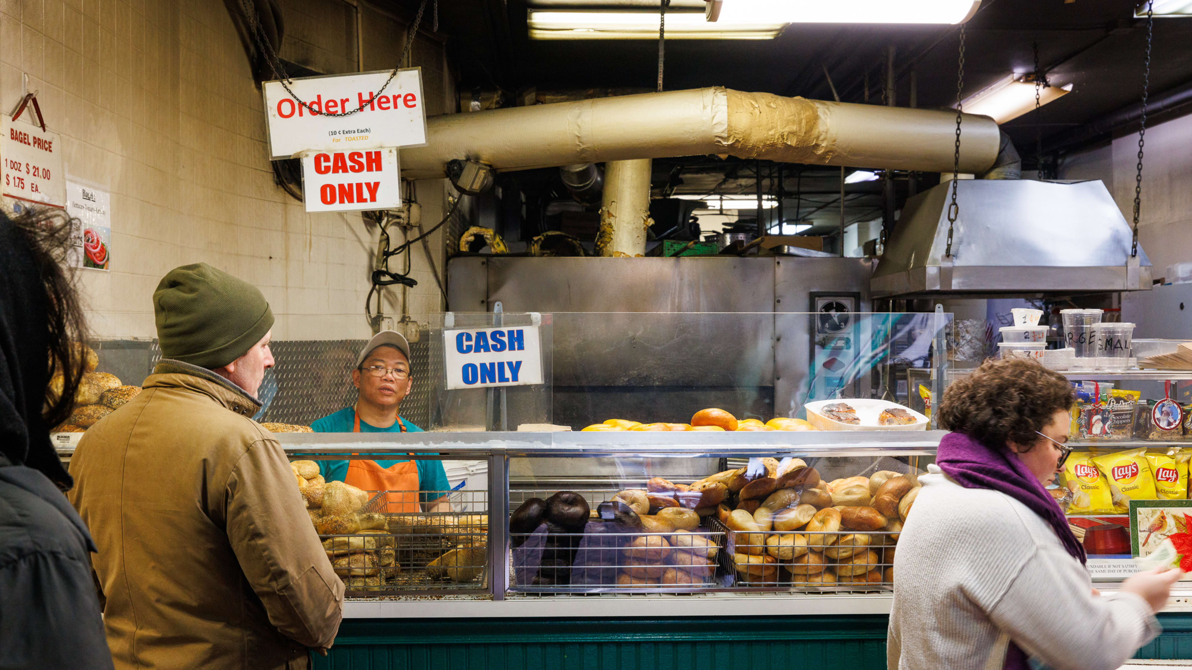 The ordering counter at Absolute Bagels.  There are bagels piled up behind the counter glass. A few people stand in line to order from an employee behind the counter.  A person pays for their order on the right.