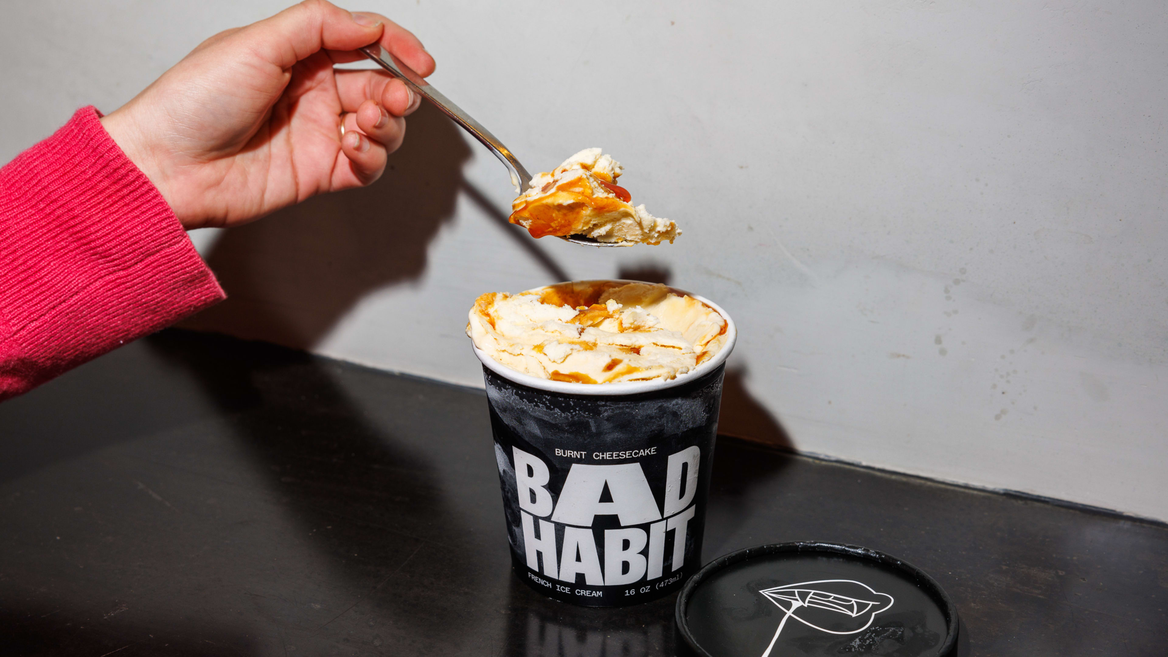 Person holding a spoonful of burnt cheesecake ice cream from a black pint with white lettering, on a black countertop.