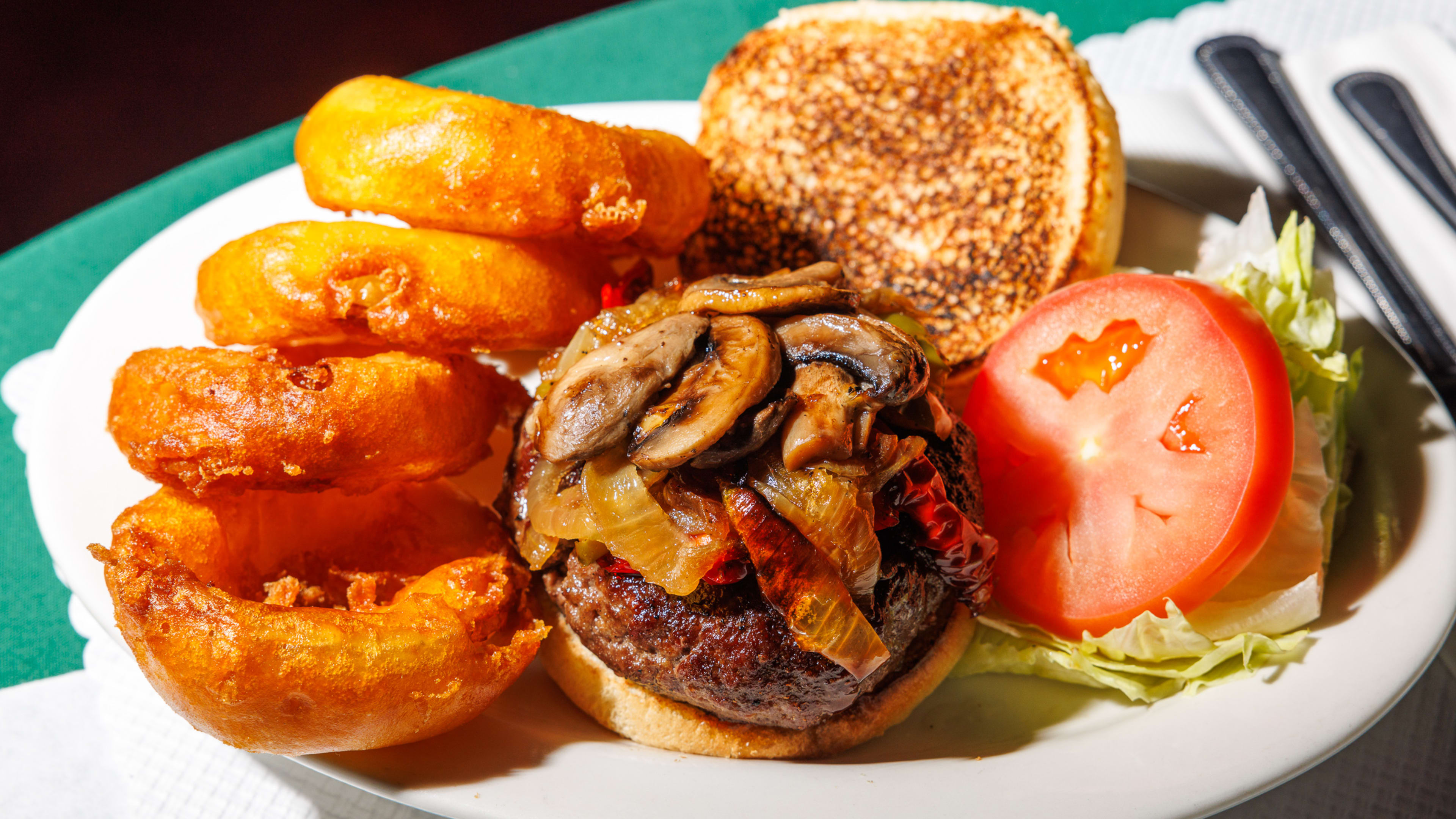 A special burger with mushrooms and onions on a plate next to a slice of tomato and lettuce and onion rings.