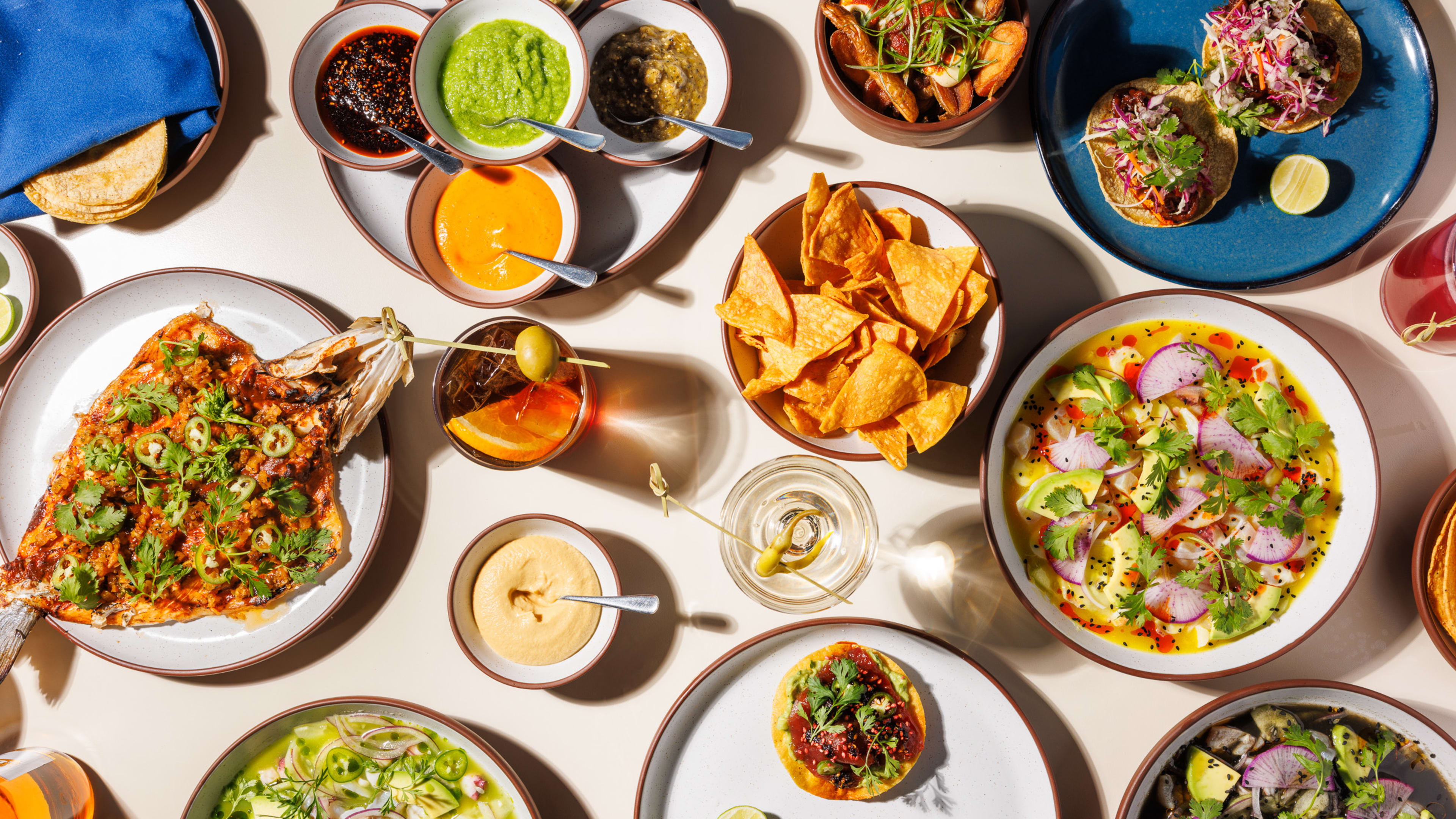 A large spread of colorful dishes including aguachile, fish, sauces, tacos, and cocktails.
