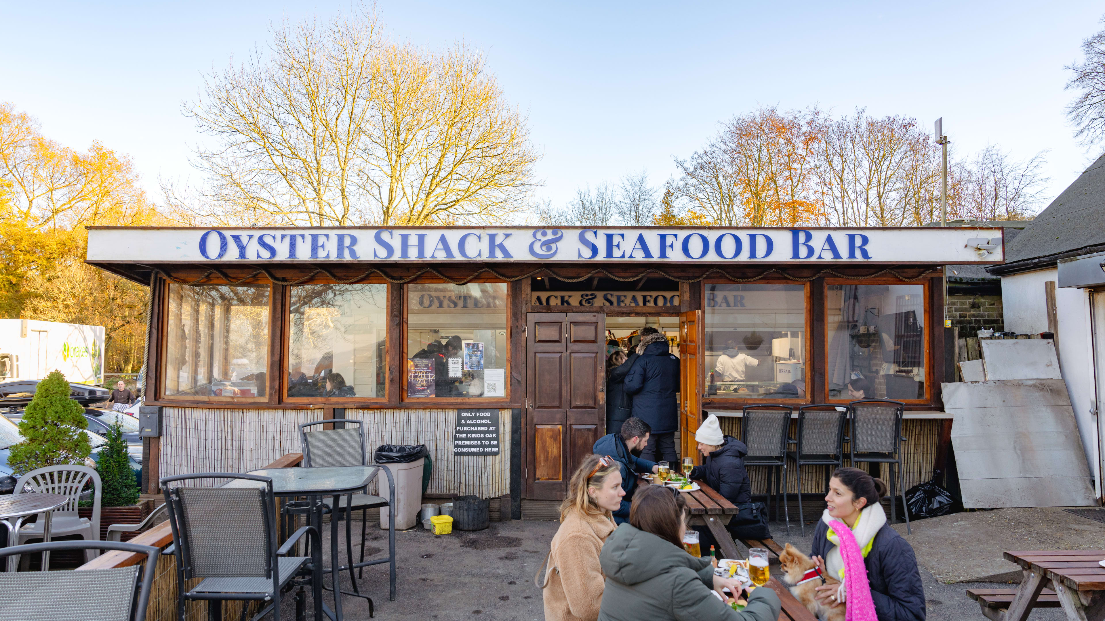 The exterior of Oyster Shack & Seafood Bar with people sitting at one of the picnic tables and people standing in line to order at the door to the seafood counter