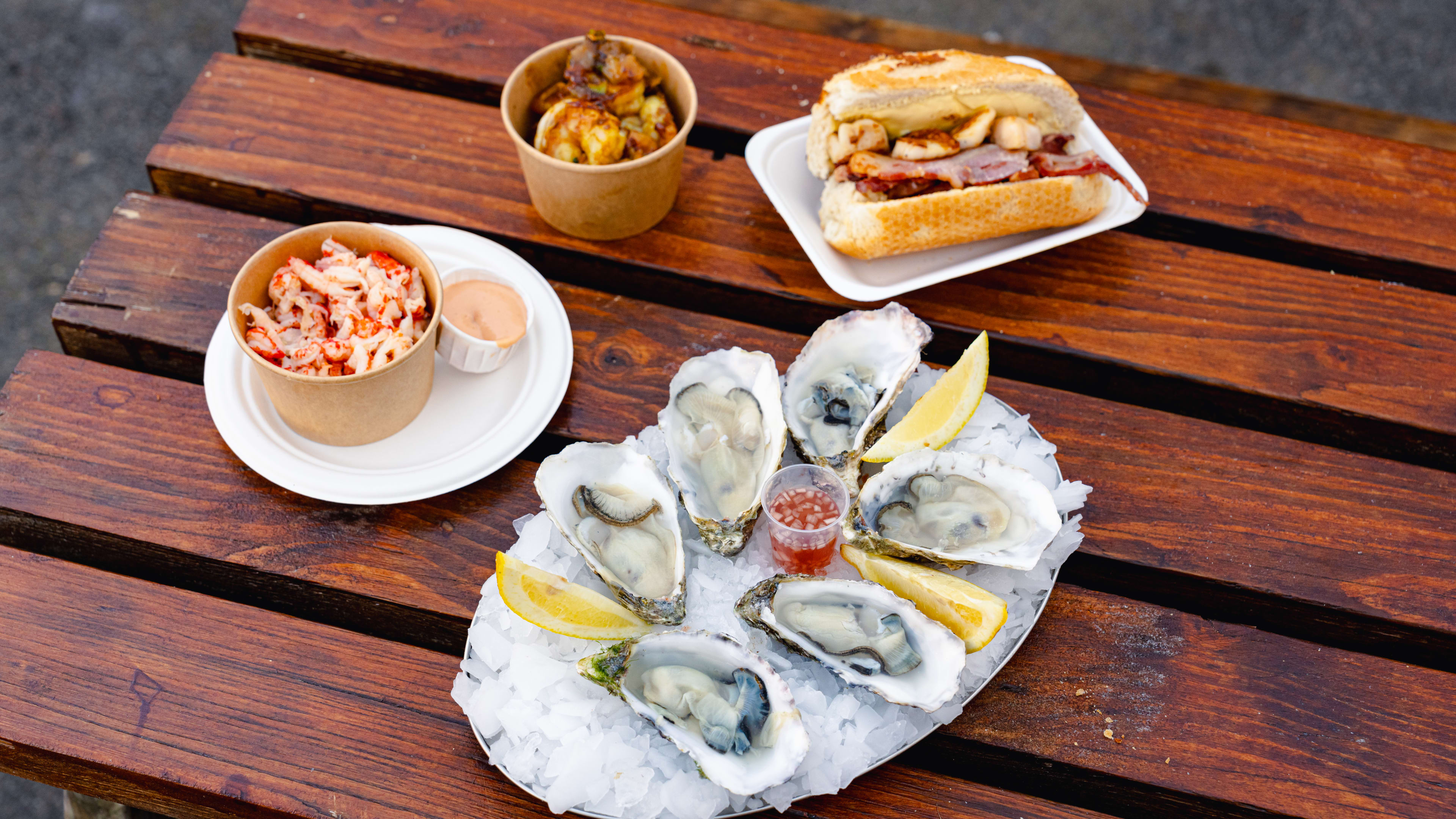 A wooden picnic table with a plate of oysters on ice, a takeaway bowl of crawfish, a takeaway bowl of prawns, and a scallop and bacon butty on a plate