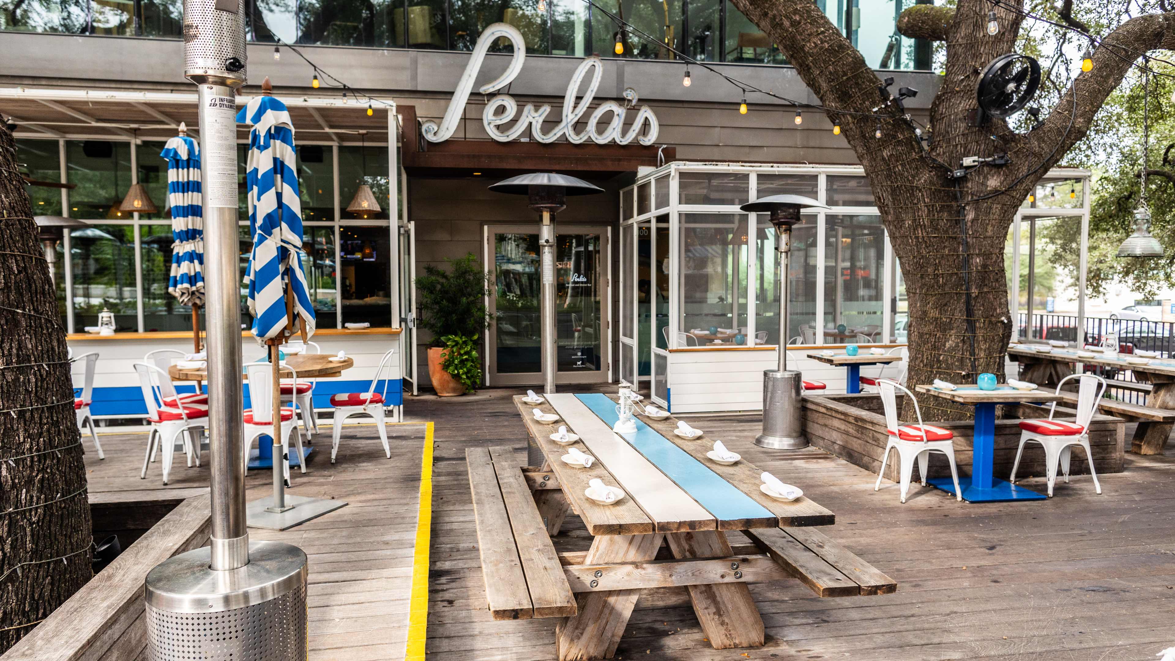 The patio at Perla's with a long wooden picnic table, round smaller tables with white chairs and string lights, heaters, and umbrellas.