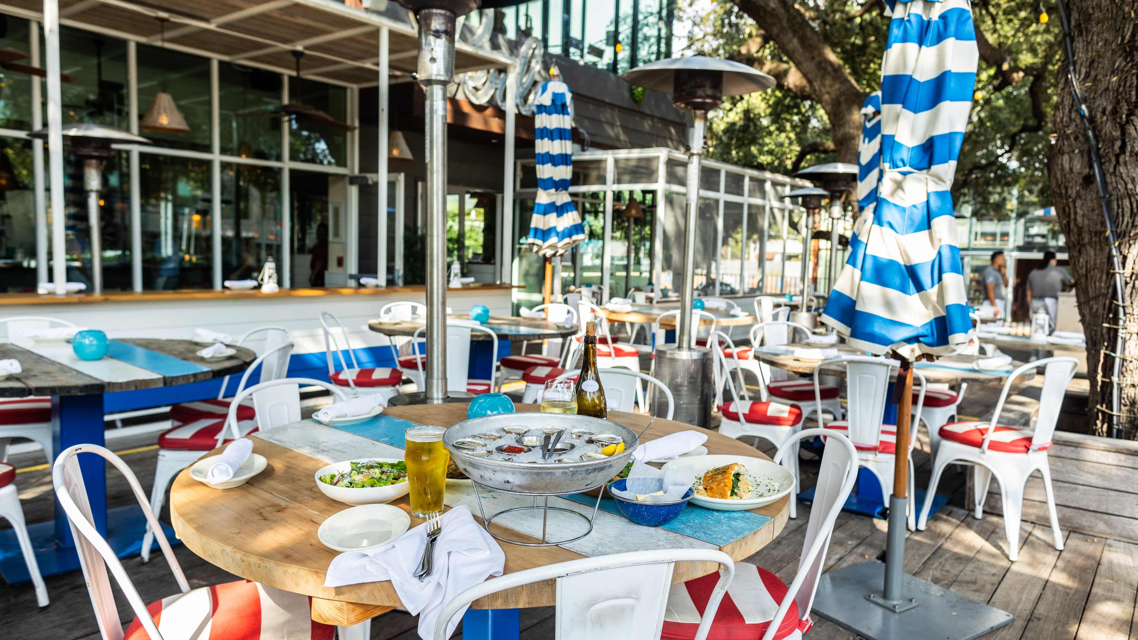 The patio at Perla's with blue and white striped umbrellas, heaters, red cushioned white chairs, and a spread of dishes on the table in the foreground.
