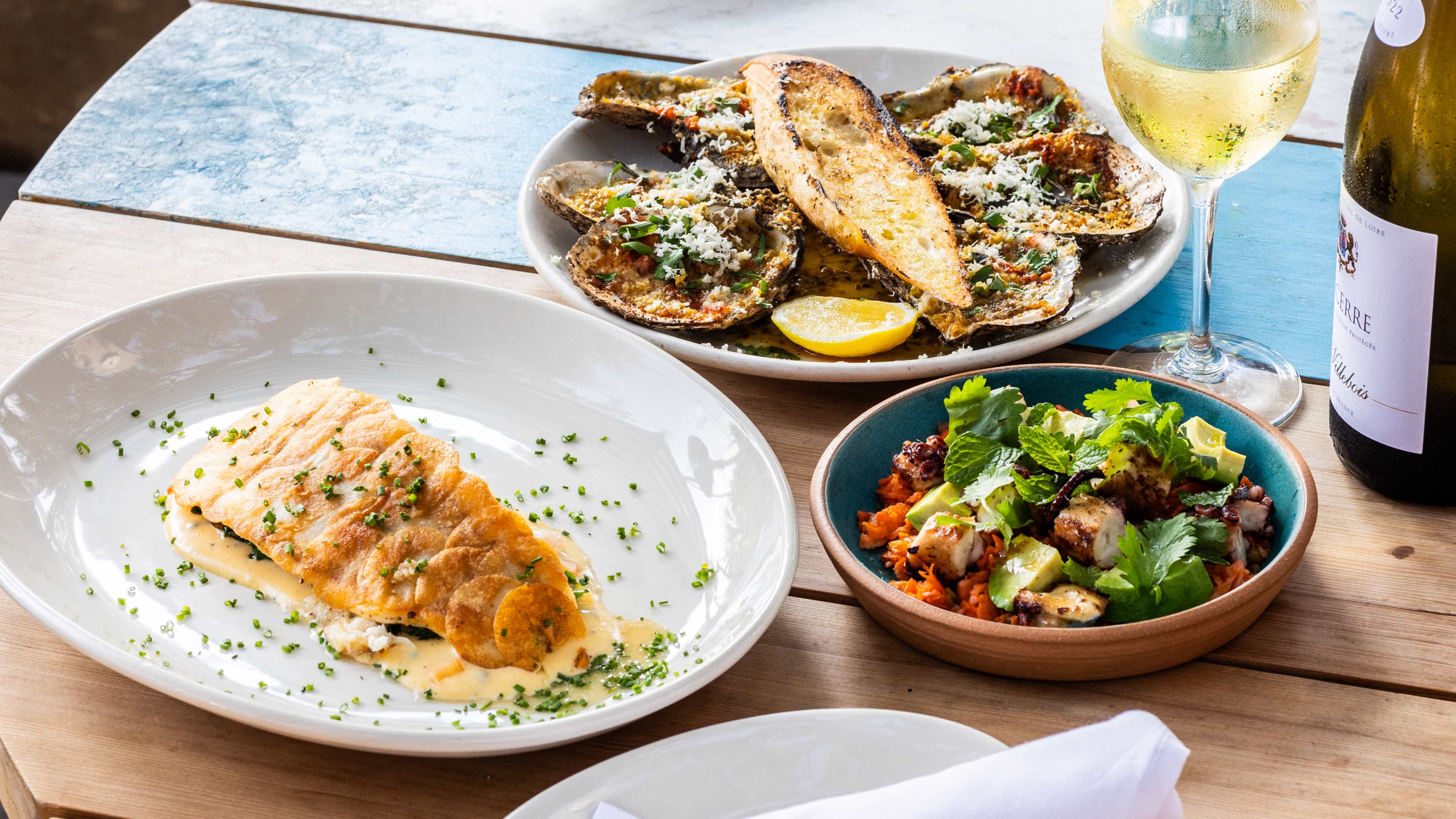 A plate of Rockfish, a blue dish with Octopus, and a plate of wood grilled oysters on a wooden round table.  There is a bottle of wine with a glass on the table as well.