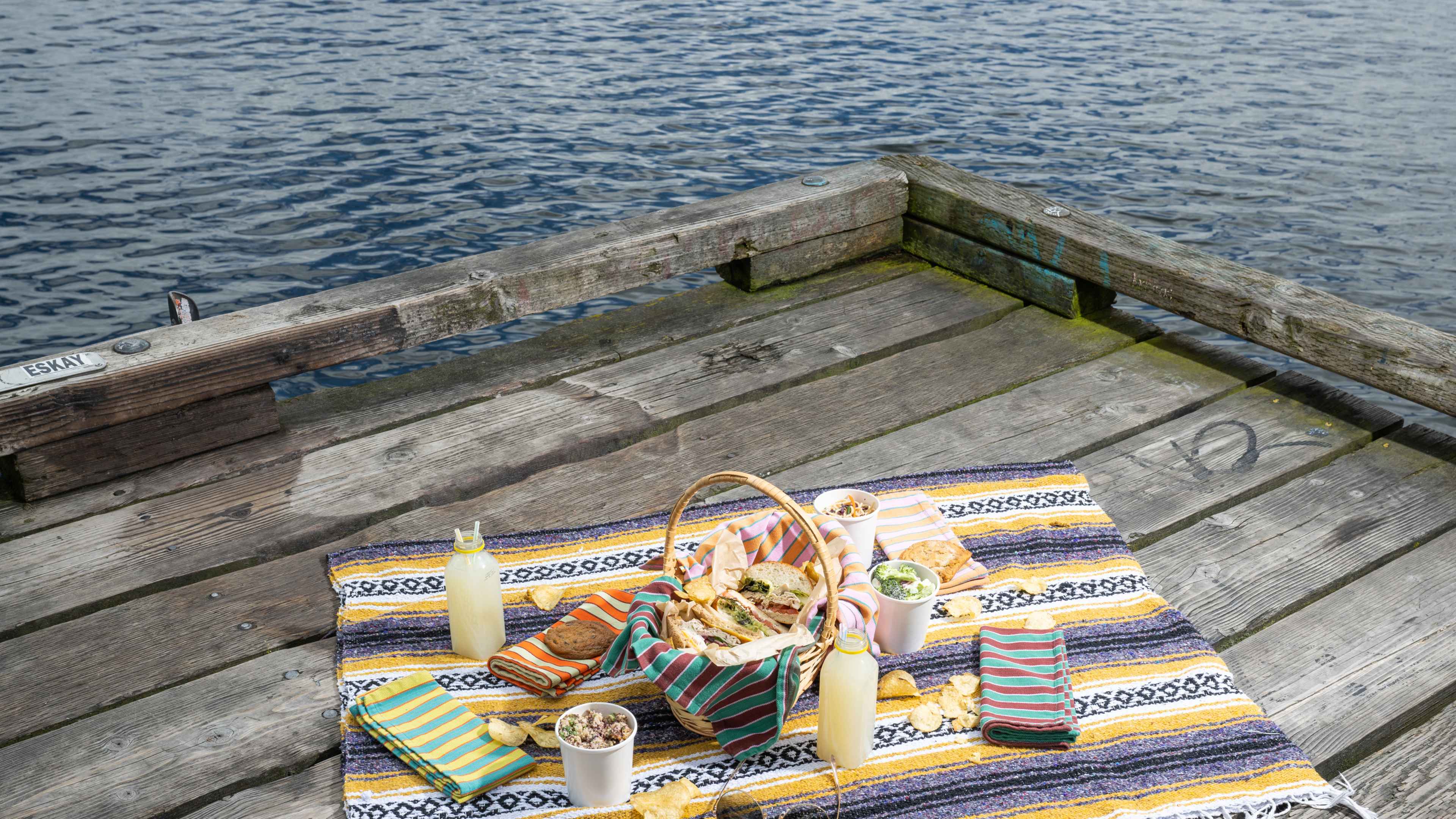 picnic blanket with drinks and sandwiches and colorful napkins on dock with water visible in background