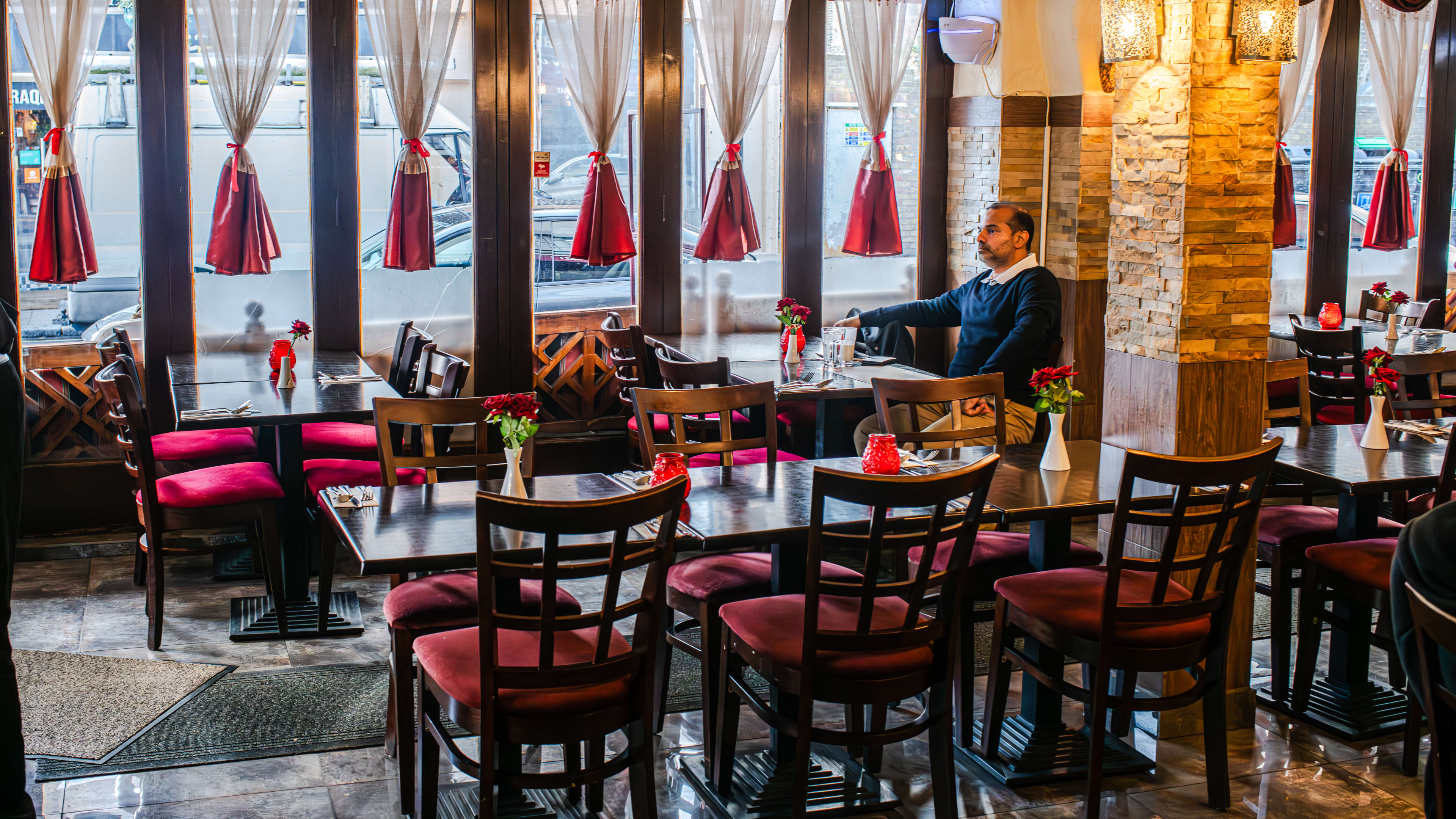 A restaurant interior with wooden tables, wooden red-cushioned chairs and floor-to-ceiling windows in the background.