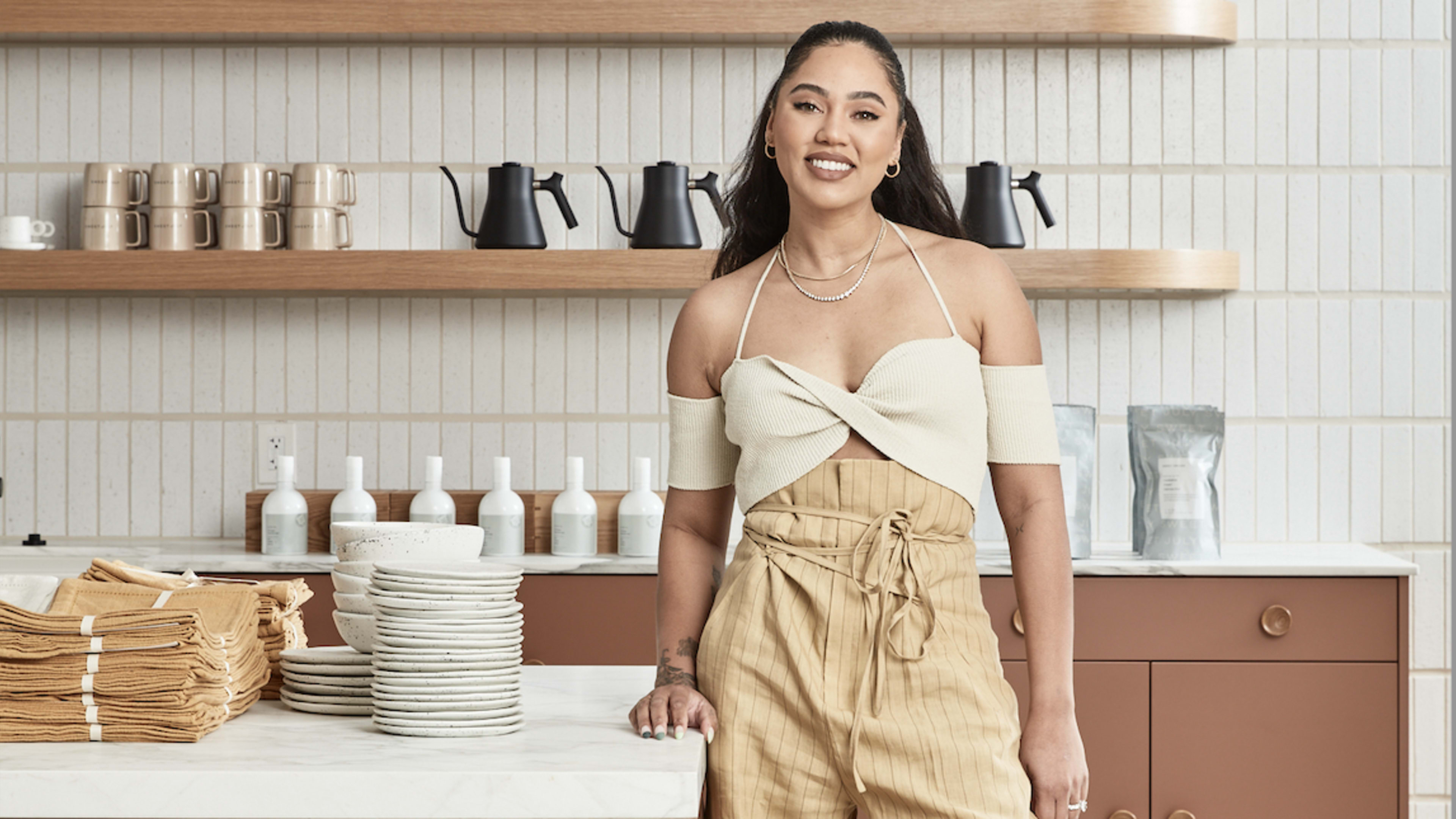 a woman stands in front of shelves containing kettles, mugs, and more