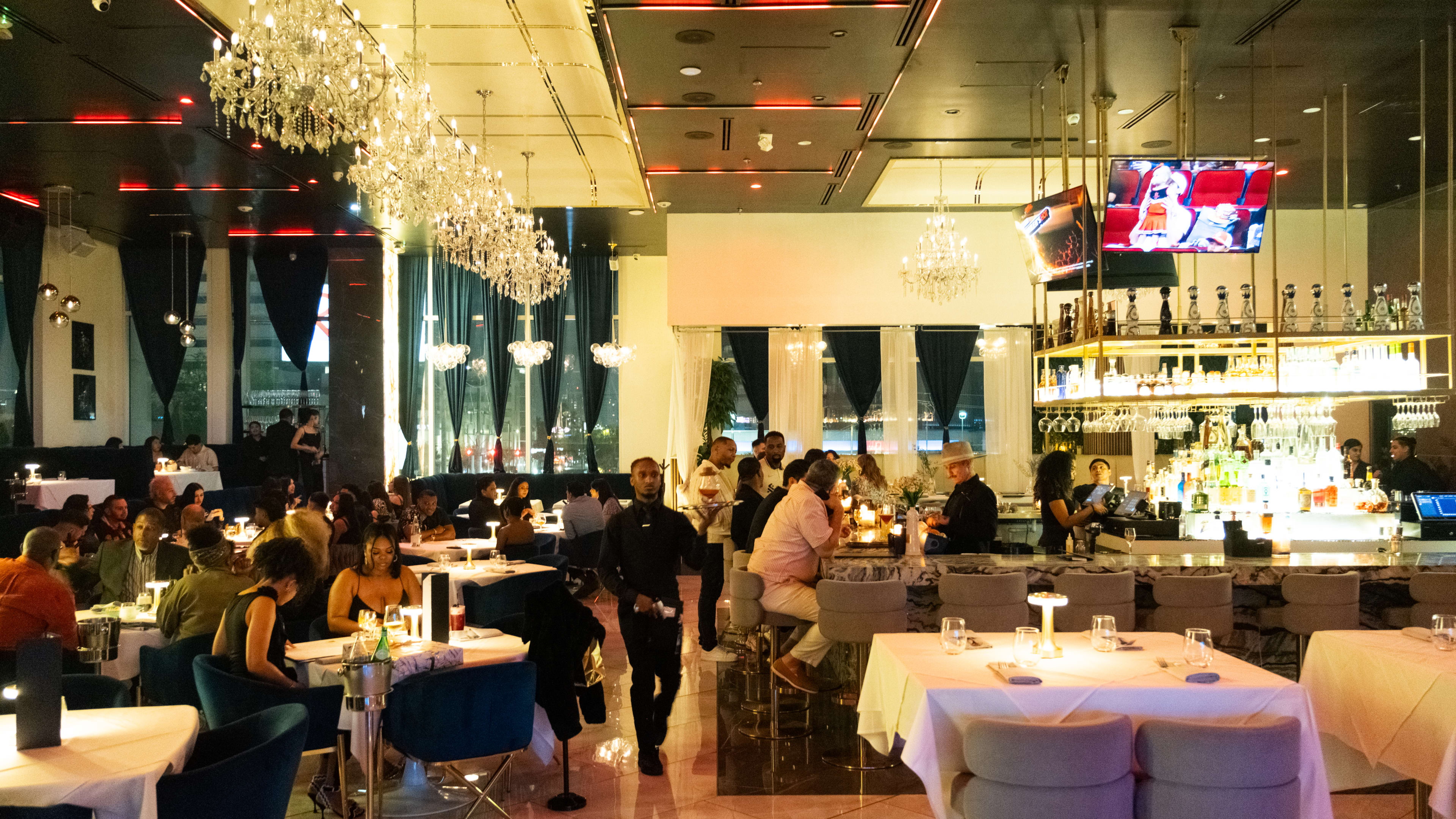 depicts interior of se7en, with white tables, a large bar, chandeliers, and blue half booths