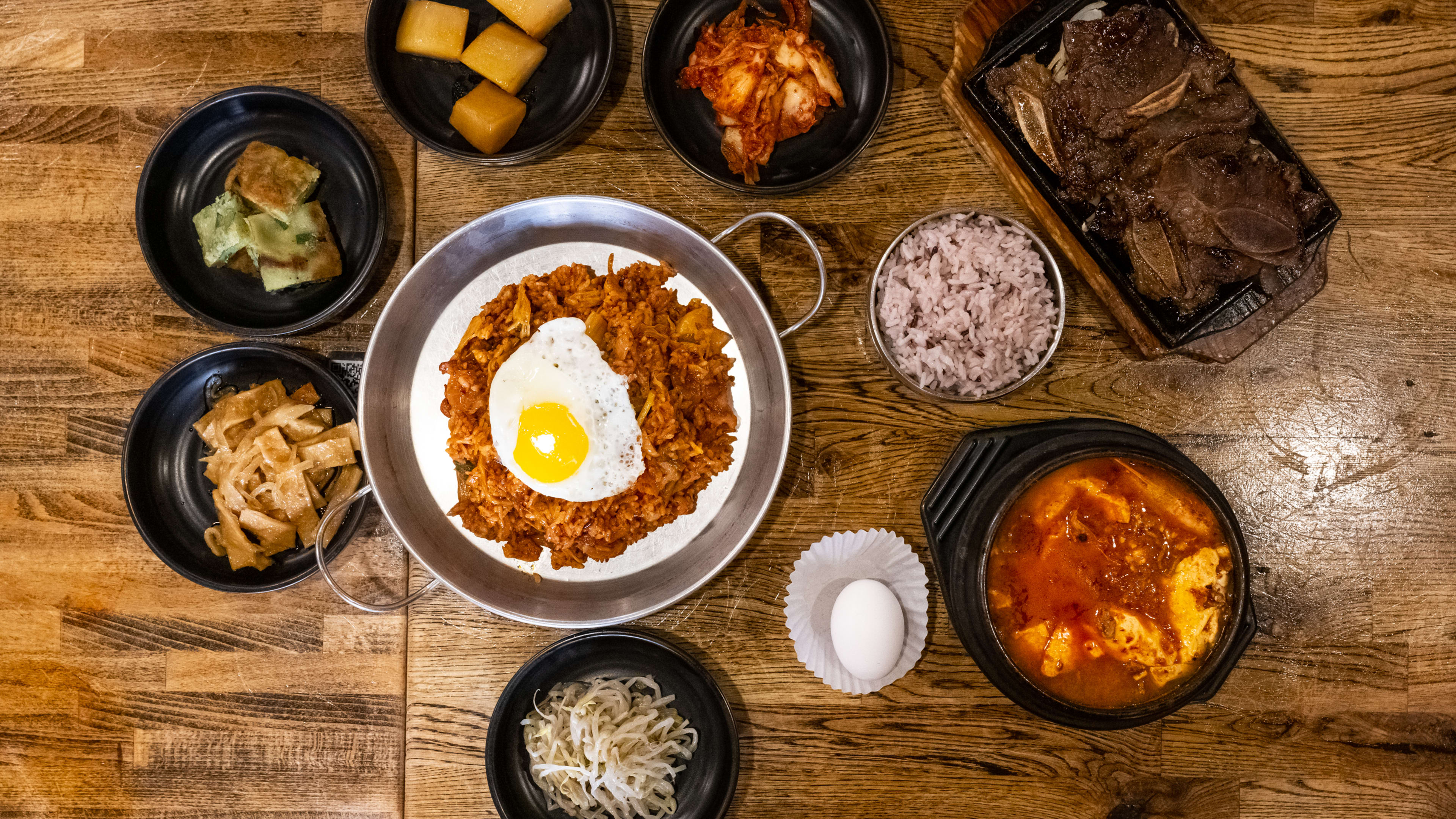 tabletop with Korean dishes on it, like kimchi fried rice topped with an egg sunny side up, kalbi short ribs, tofu stew, and various banchan.