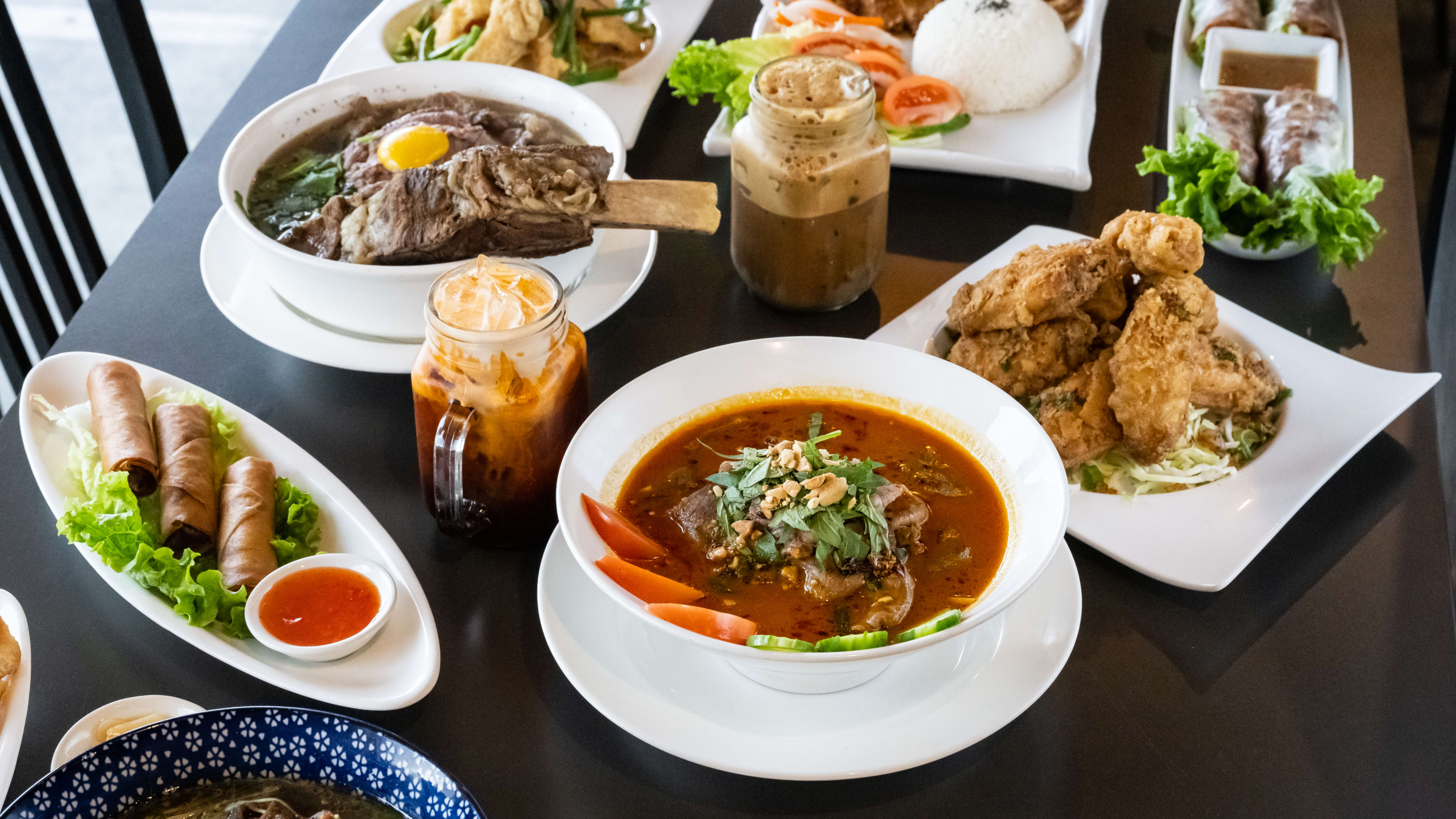 Spread of dishes like pho, egg rolls, Thai iced tea, Vietnamese iced coffee, rice, and chicken.