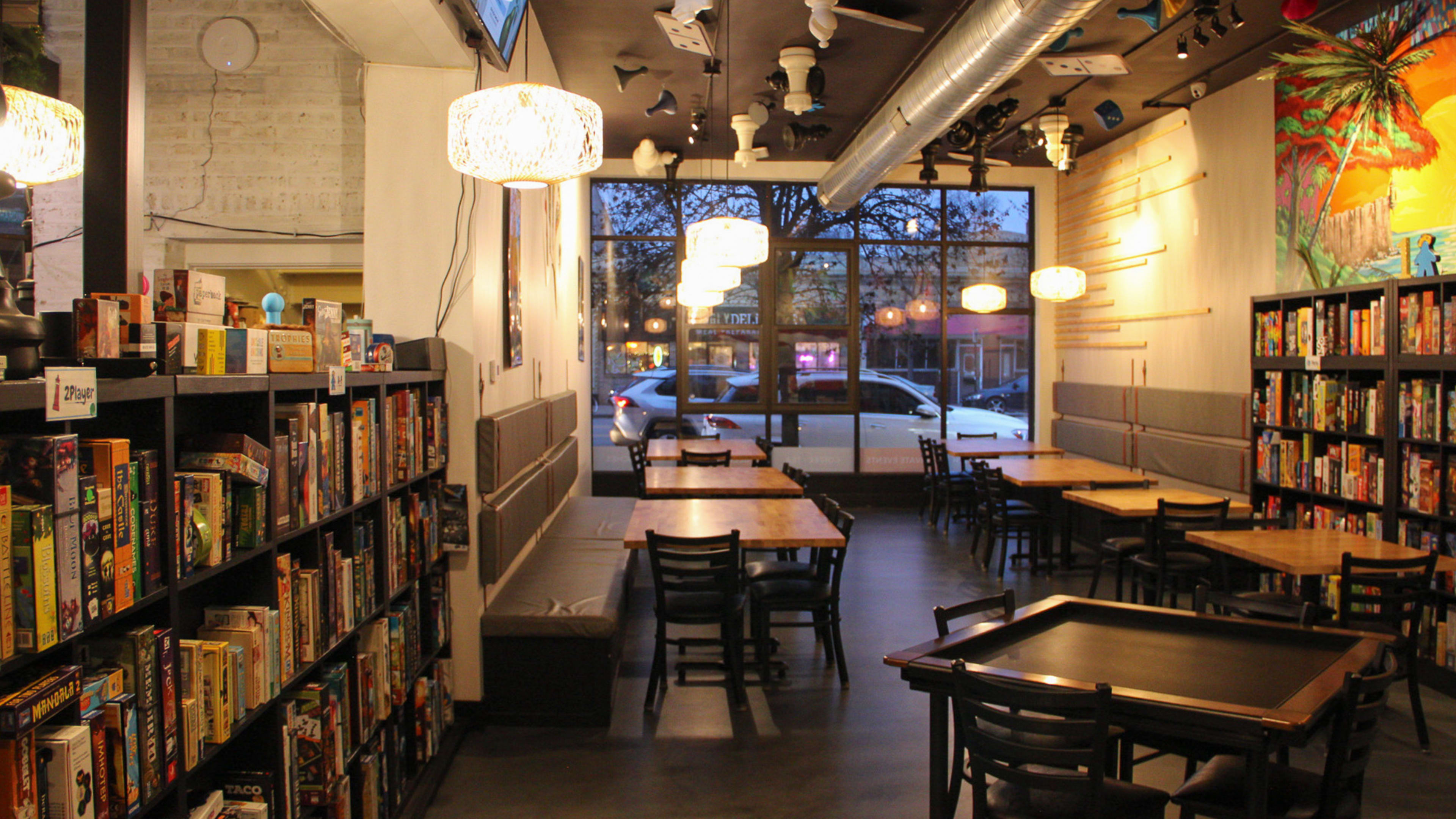 The interior of Stay and Play Game Cafe. There are tables next to large bookshelves filed with games. There is a colorful painted mural on the wall.