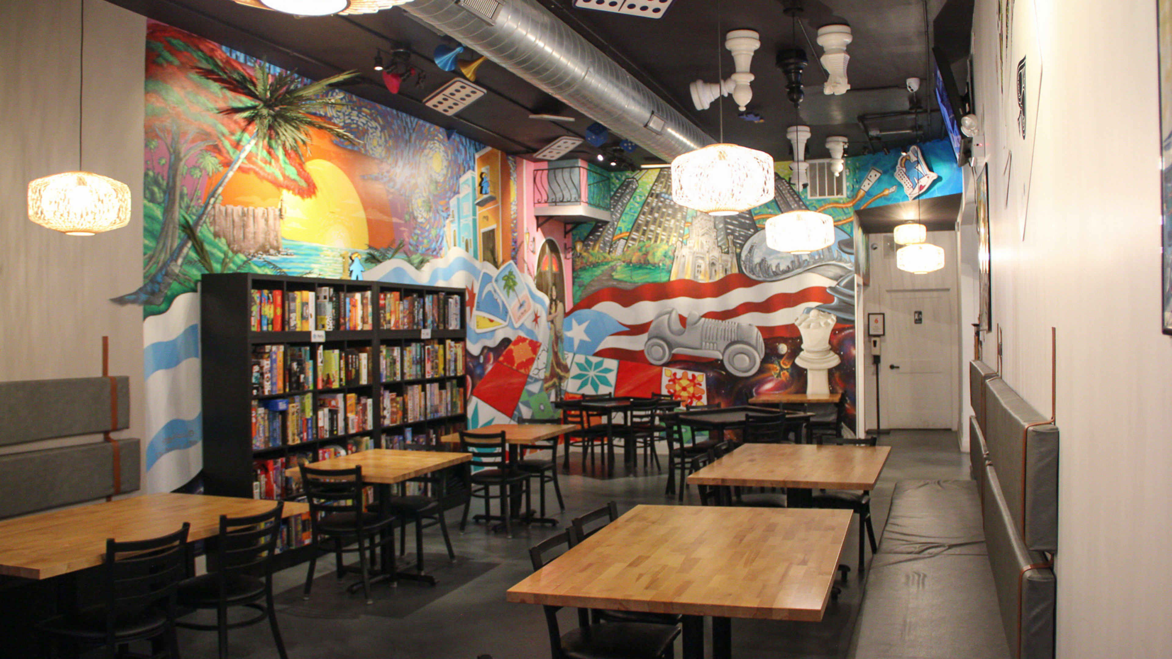 The interior of Stay and Play Game Cafe. There are tables next to large bookshelves filed with games. There is a colorful painted mural on the wall.