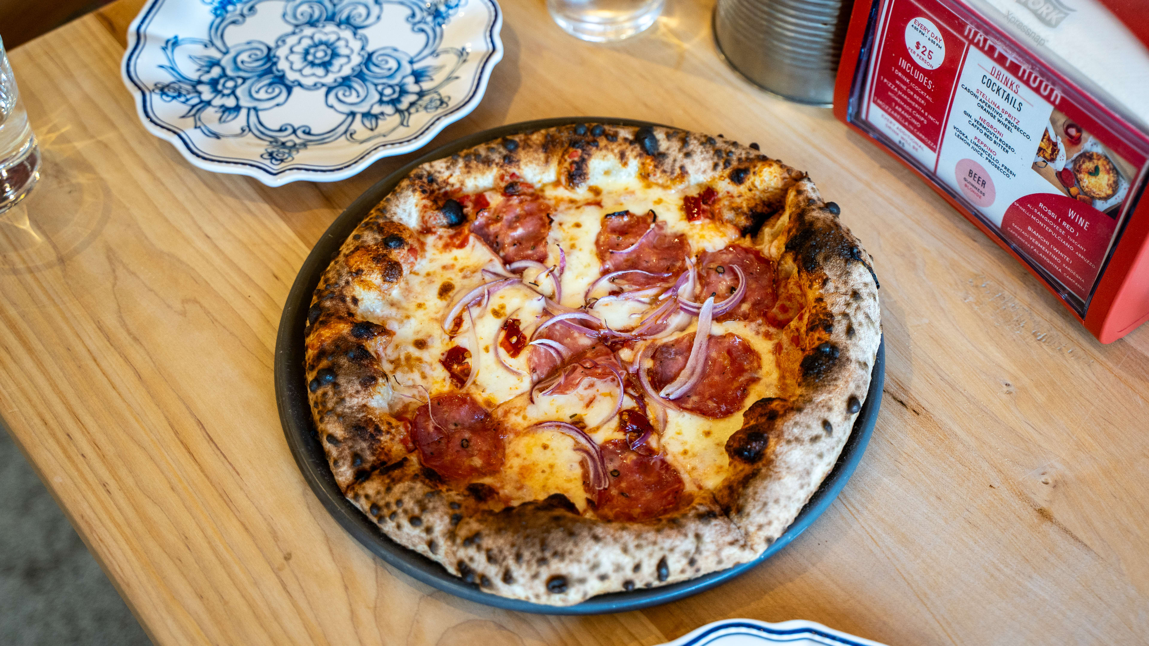 Pepperoni pizza with charred crust and red onions on table with two china plates.