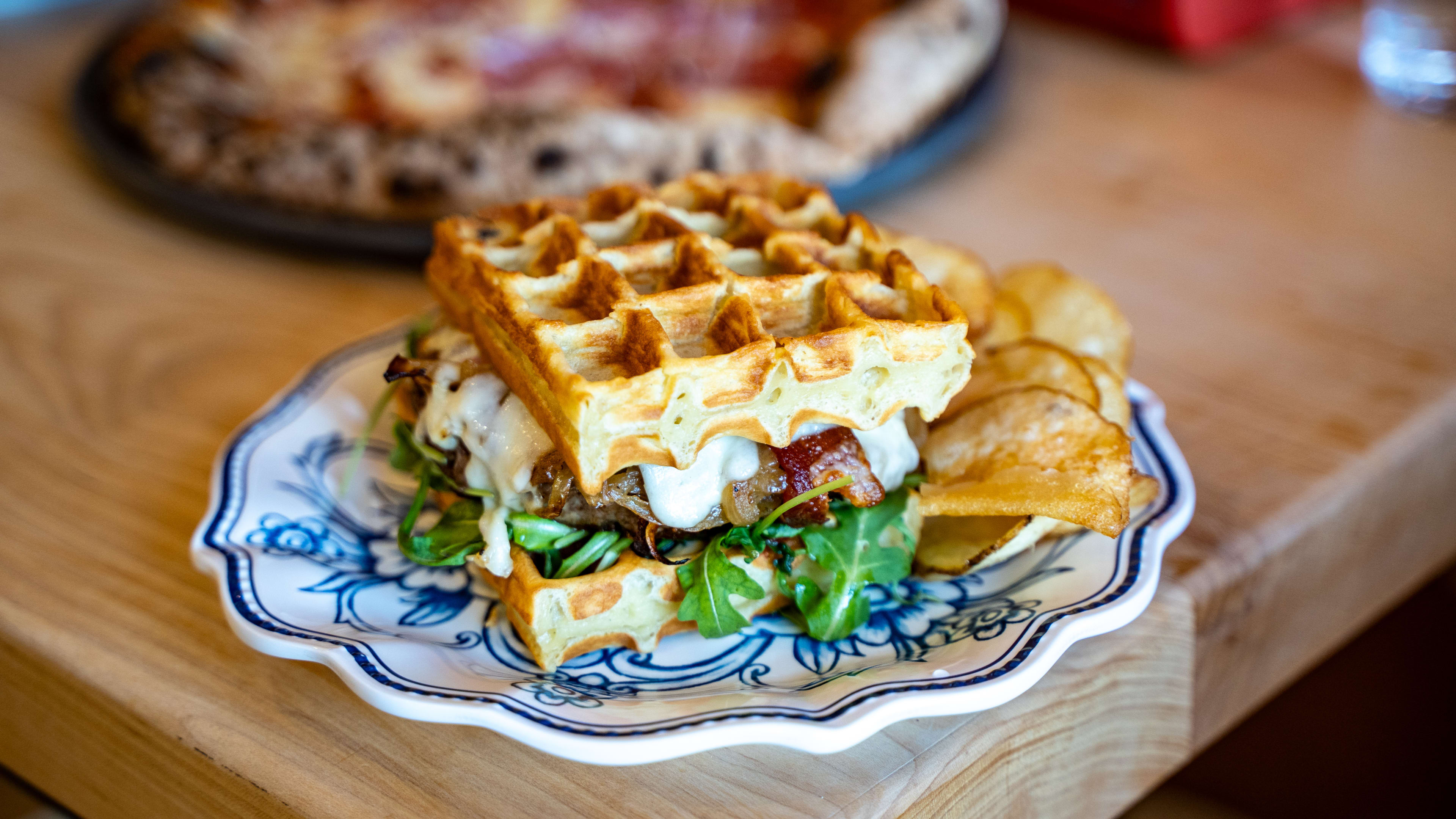 Burger between two waffles topped with arugula, bacon, and cheese, and served next to chips on a china plate.