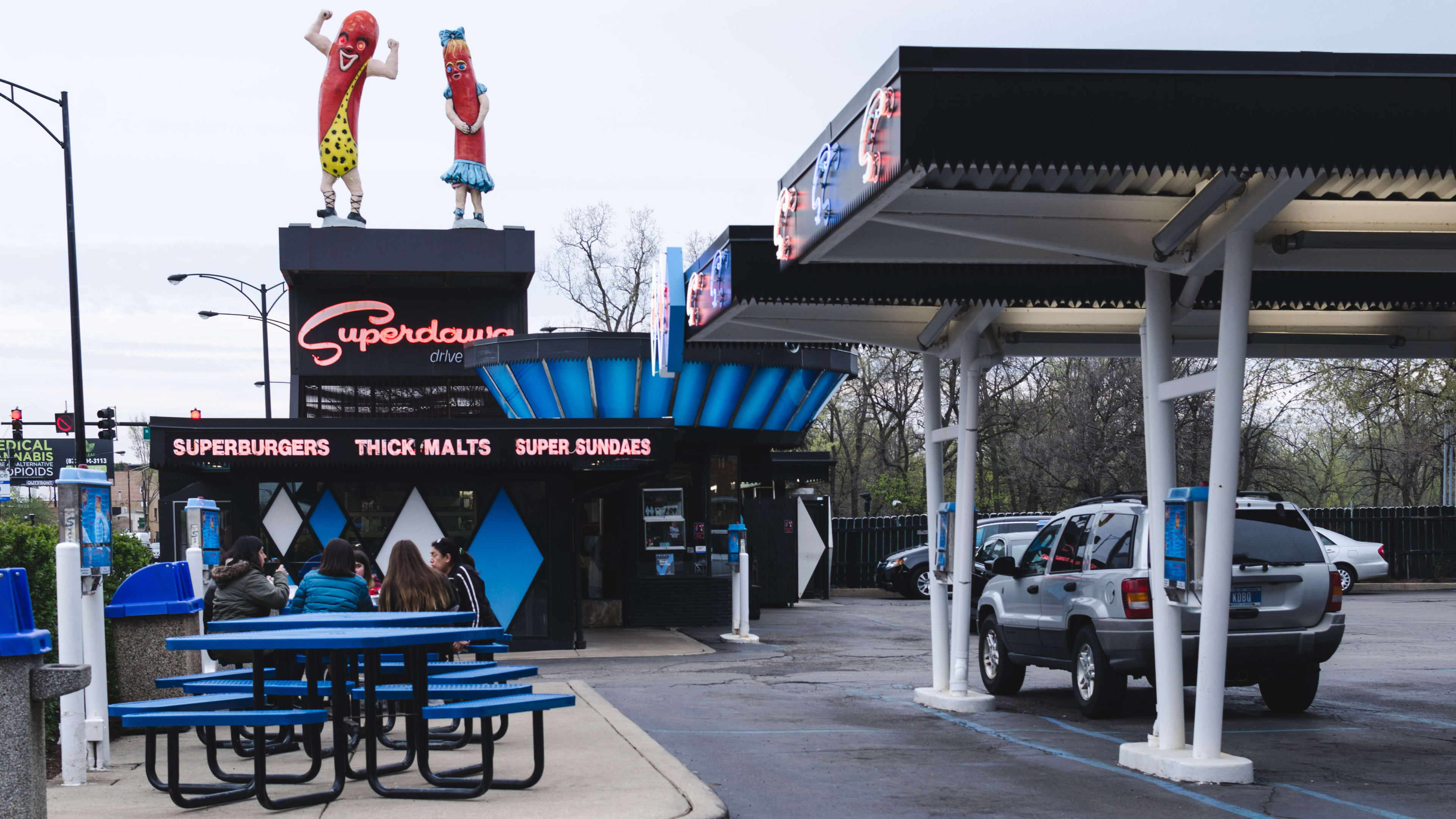 The exterior of the Superdawg drive-thu with a covered area to park and blue metal picnic tables.