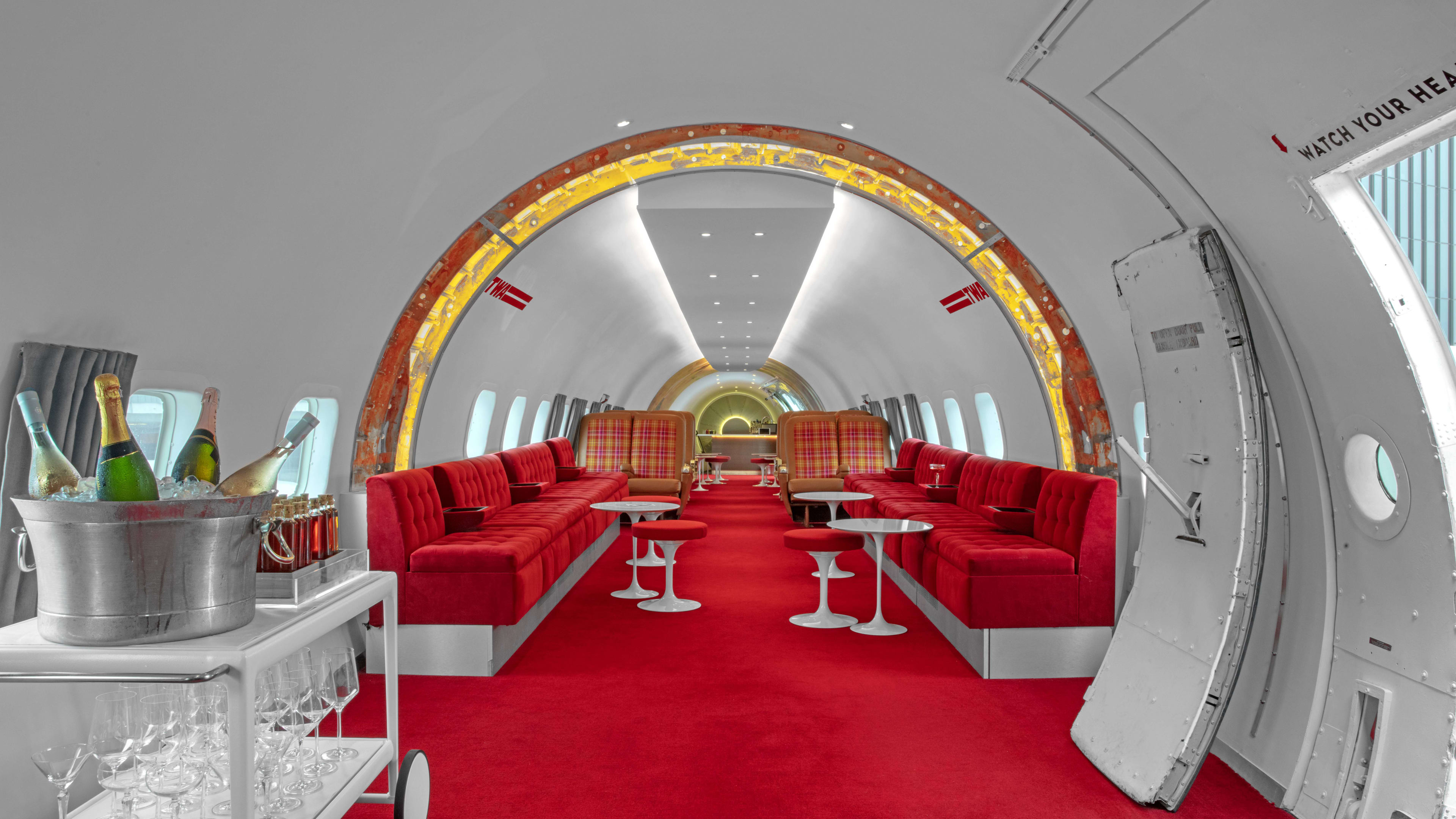 The converted retro airplane interior of Connie bar. The floor is covered in red carpet and there are long red booths, with small white tables and accompanying small stools. There are also airplane seats in the back. There is a white drink cart in the foreground.