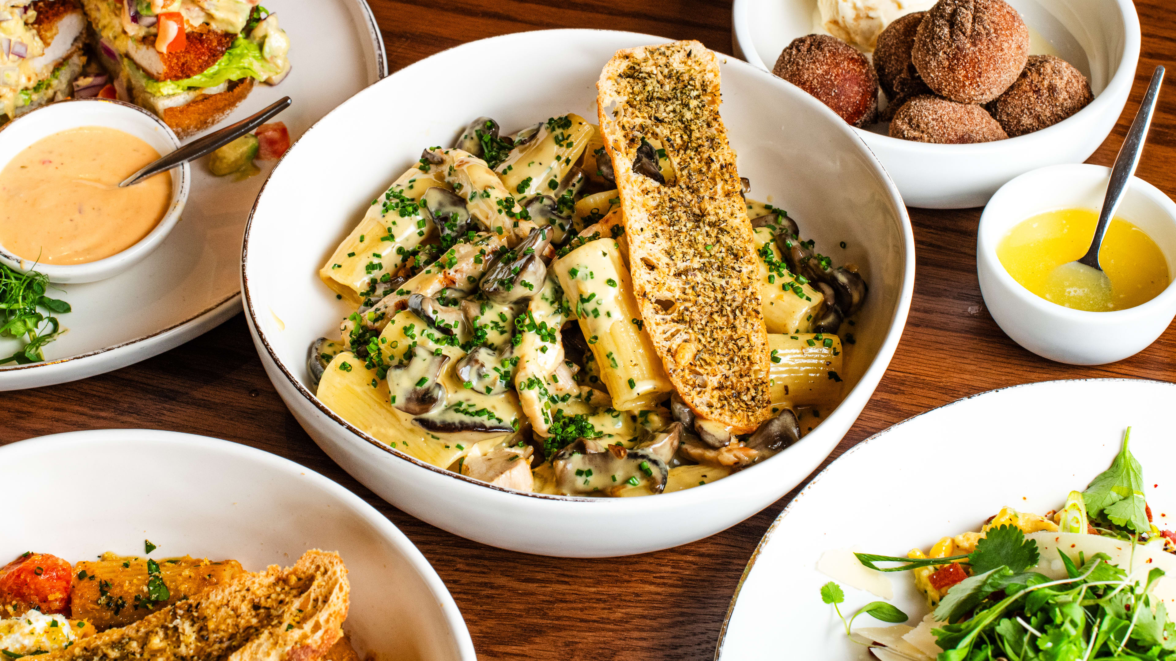 Various dishes from Tashas sit on a wooden table. Savva's Chicken Pasta is in the center.