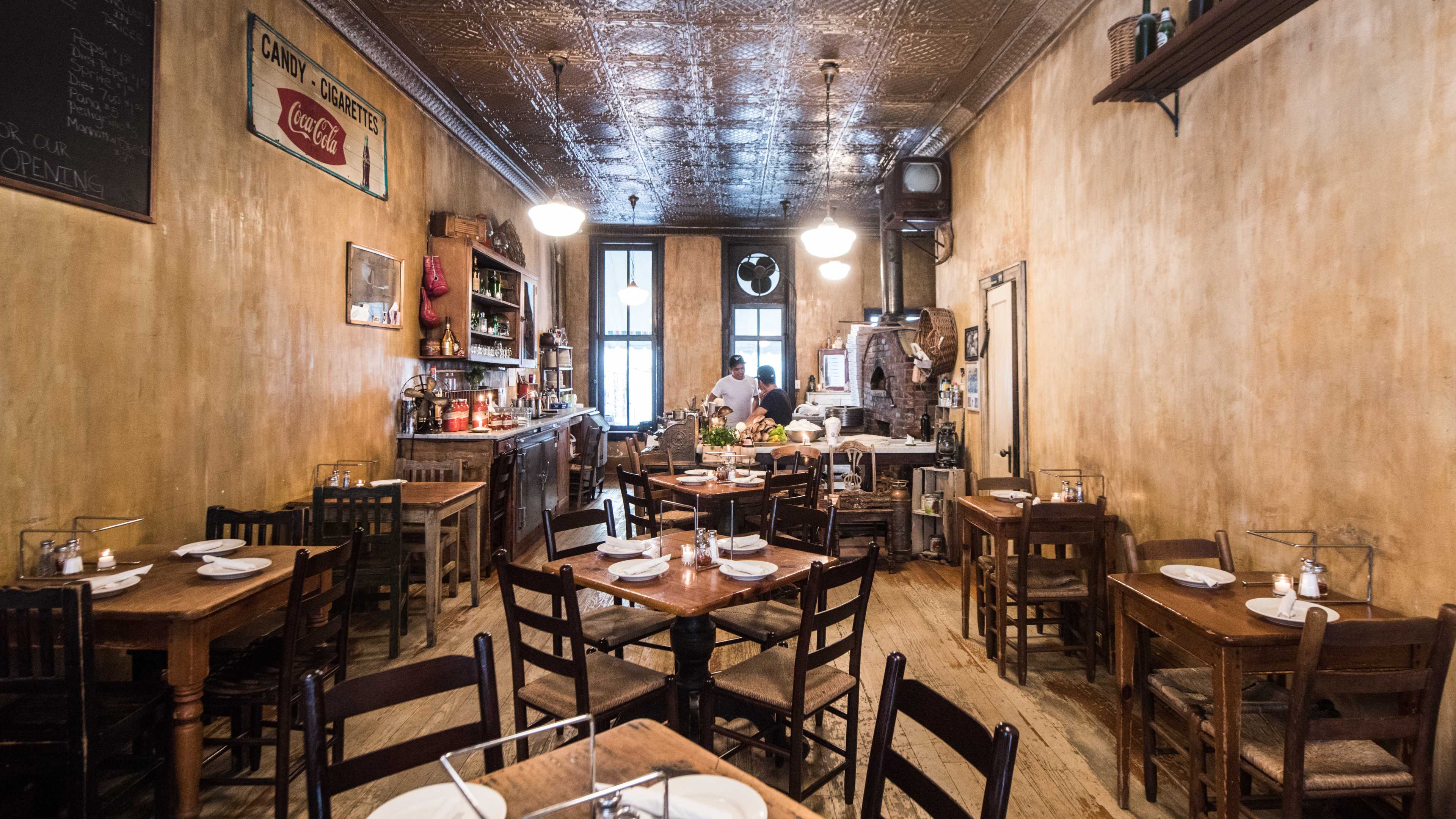 The interior of Lucali with venetian plaster-esque paint on the walls, vintage art, worn wood floors, dark wooden chairs and tables, a few shelves, and tin ceiling tiles.