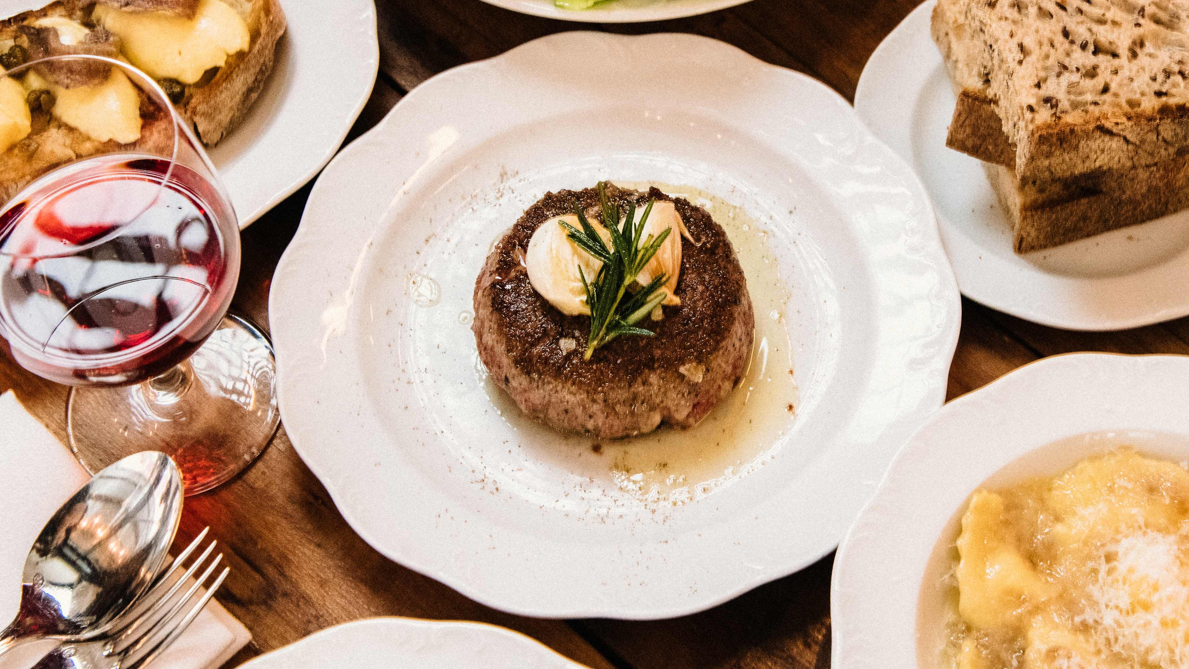 The circular steak from Via Carota, served on a white, scalloped plate with garlic and rosemary garnish on top. There are other dishes surrounding the steak as well as a glass of red wine.