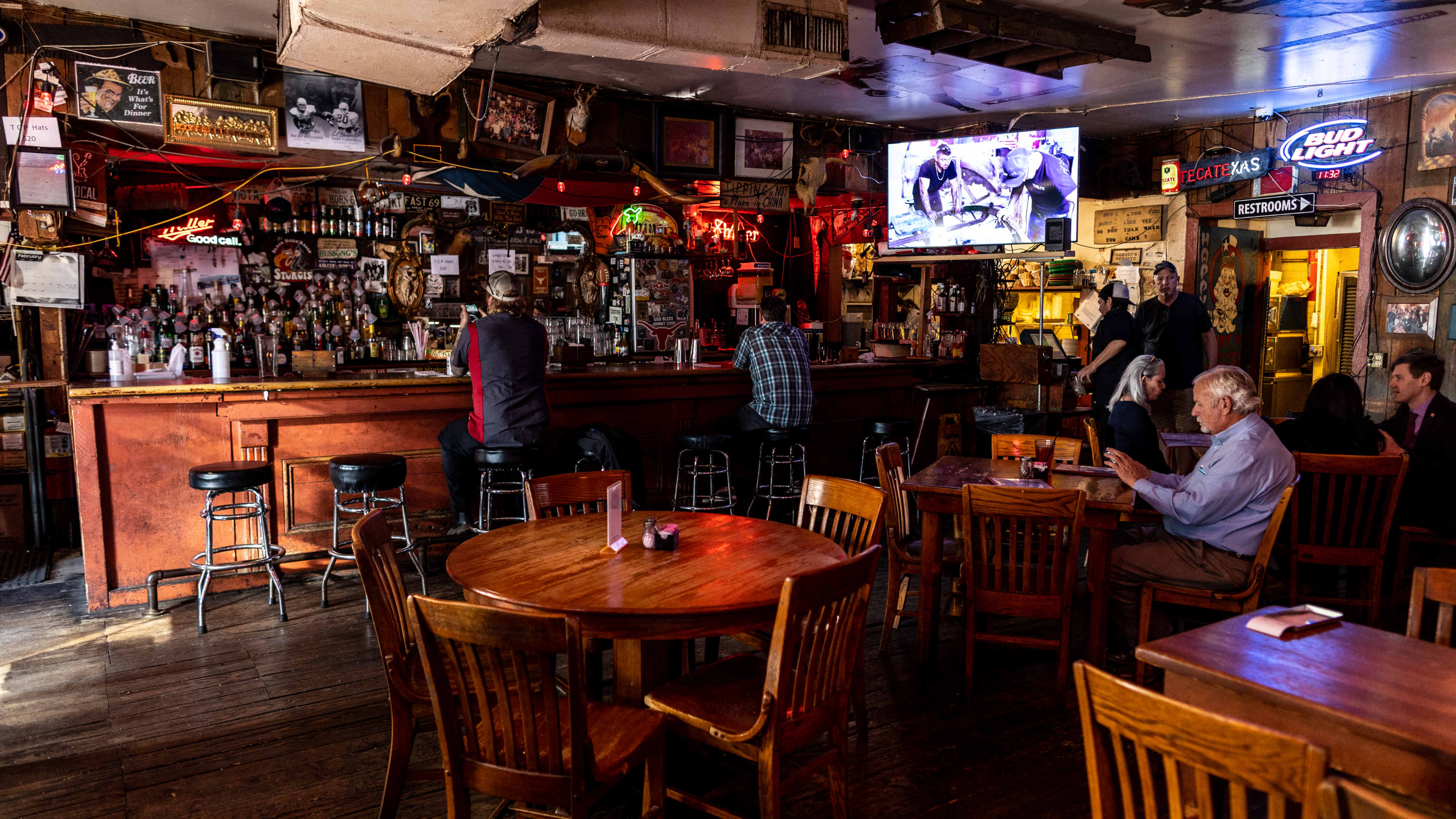 The interior of Texas Chili Parlor. There are people sitting at a wooden bar, and wooden tables and chairs.  A TV hung above the bar is playing sports.